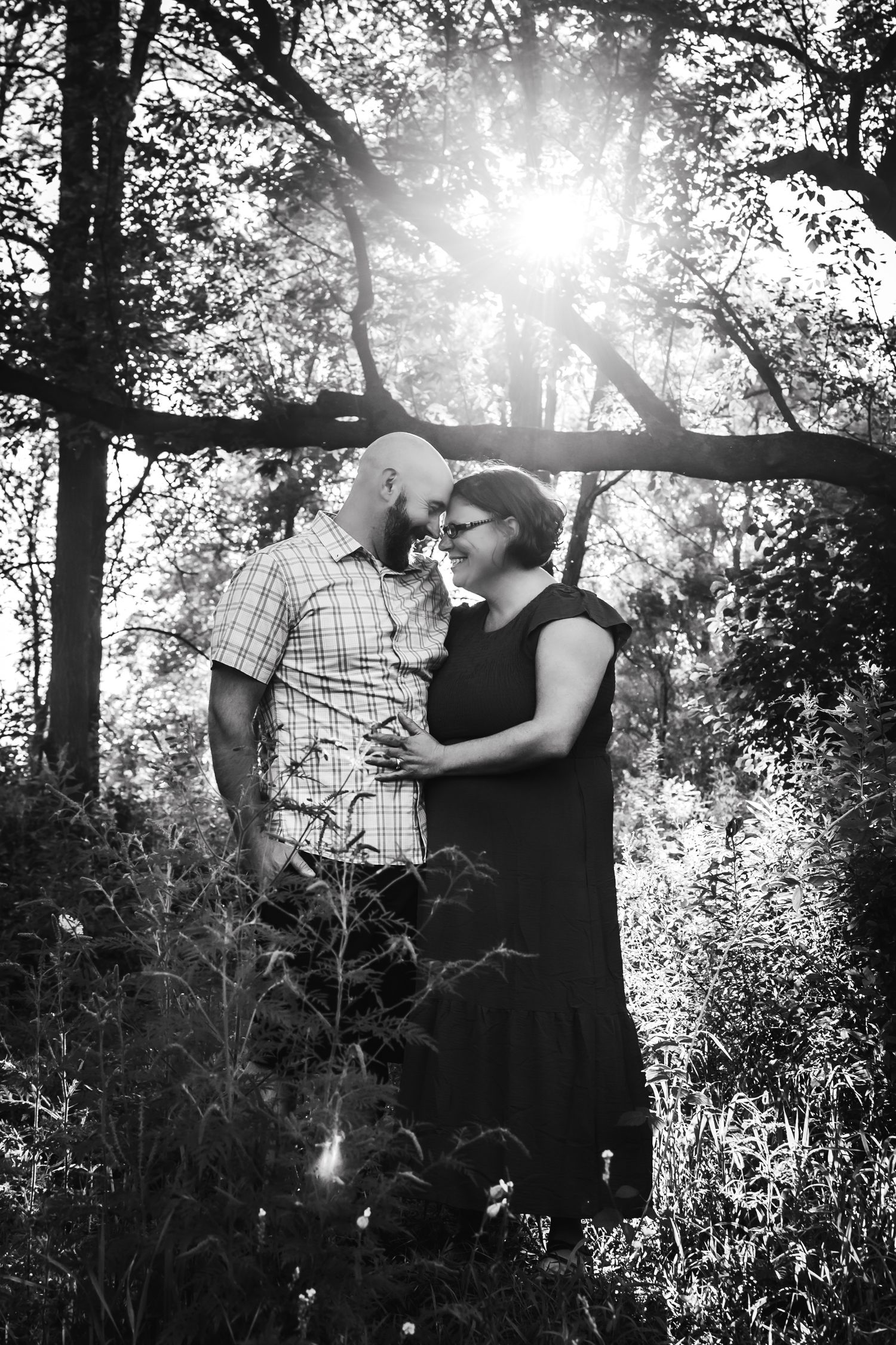 Artistic black and white couple's portrait with sunburst through trees in background.