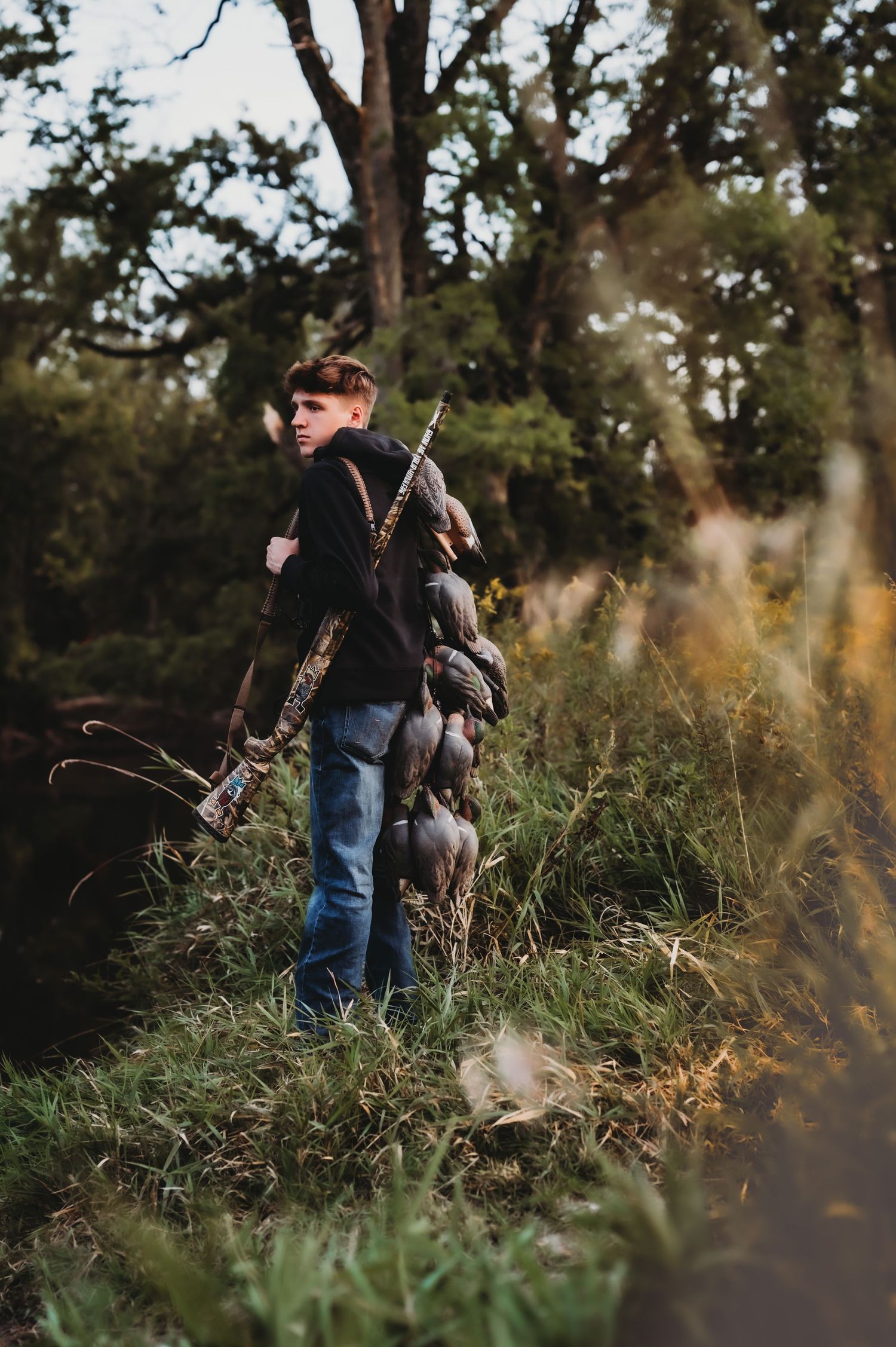 Person in casual wear holding a rifle while standing in tall grass near trees.