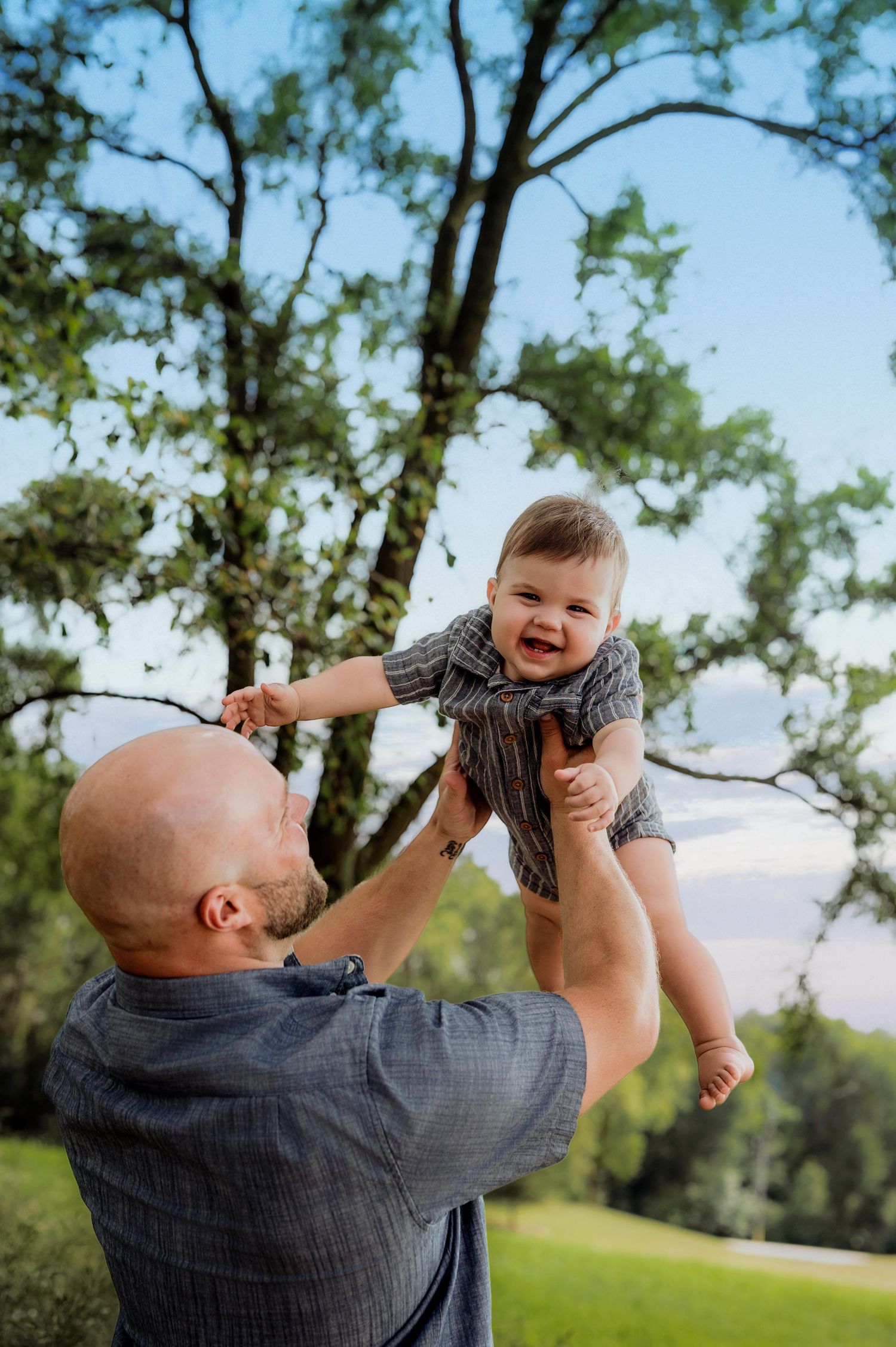 Bald father joyfully lifting laughing baby outdoors under green trees on a sunny day.