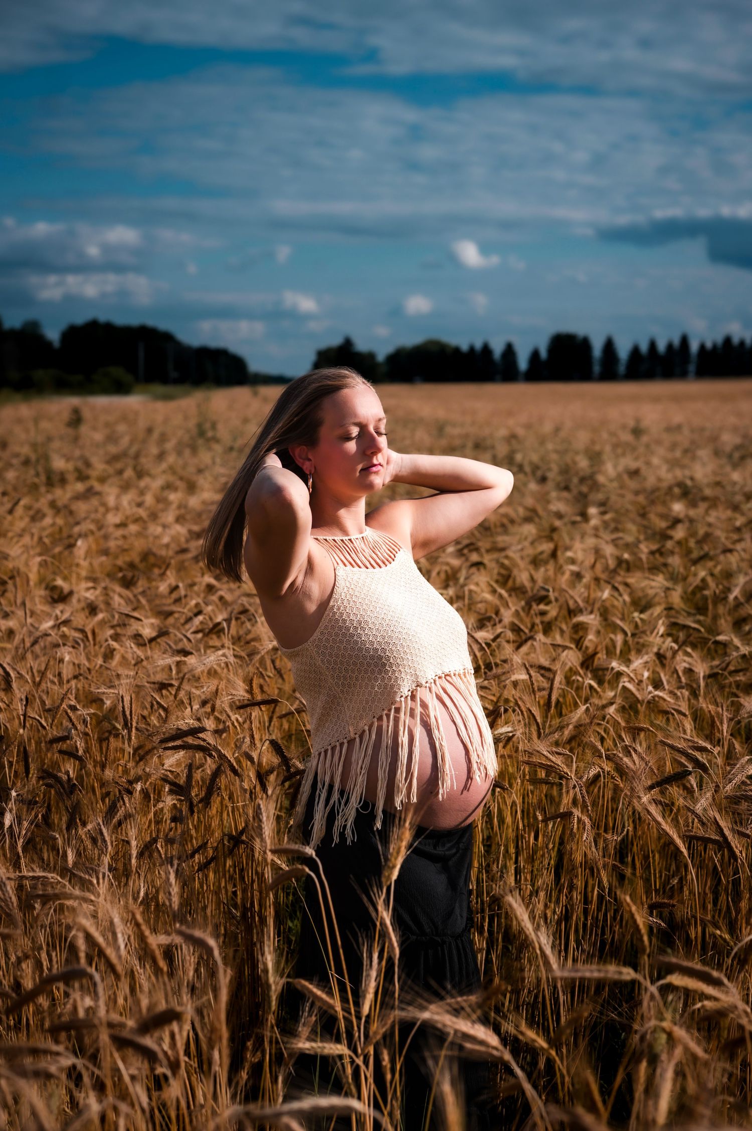 Pregnant woman in crochet top standing in golden wheat field under blue sky.