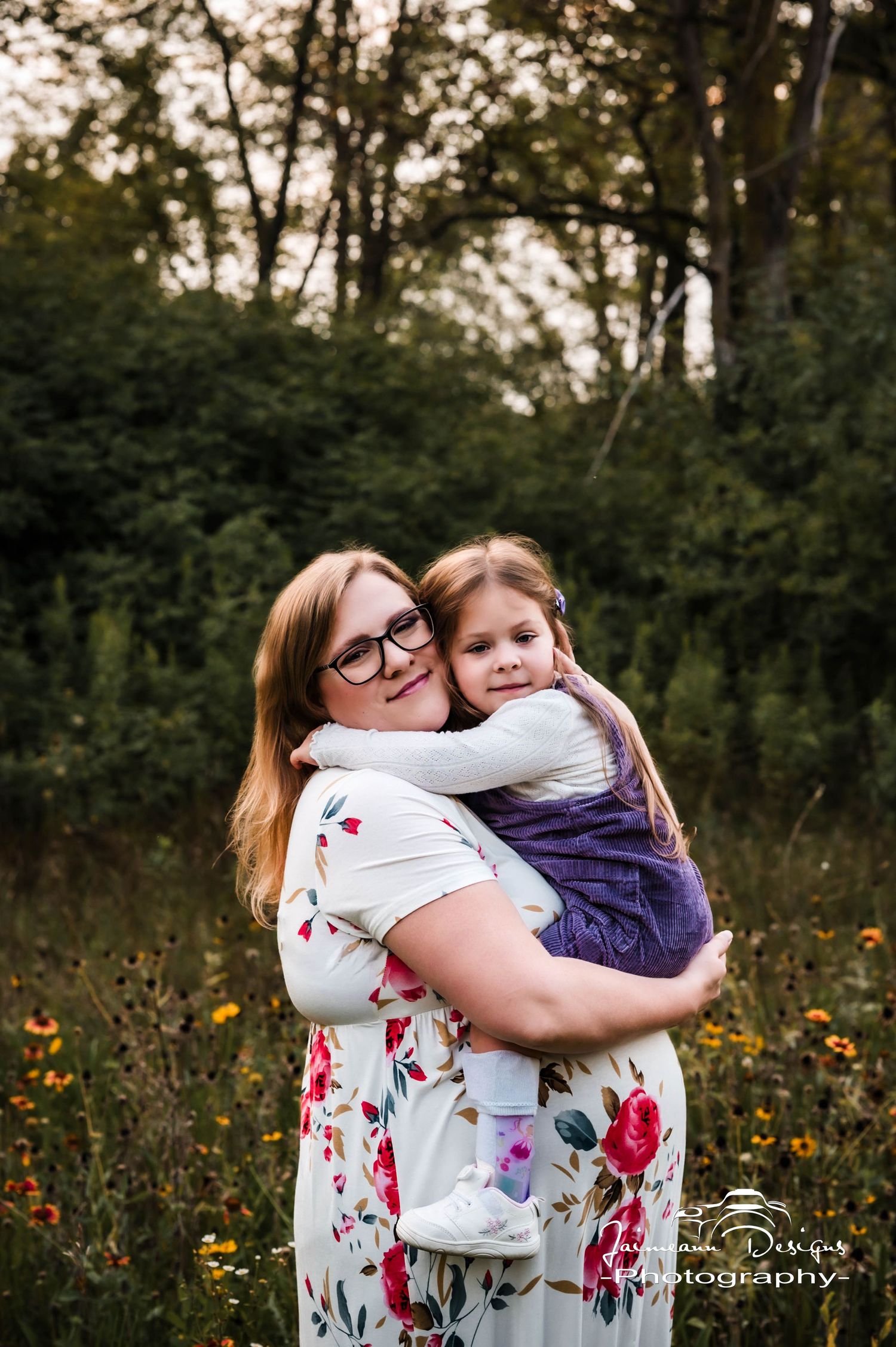 A mother in a floral dress hugs her young daughter in a sunlit meadow with lush green trees in the background.