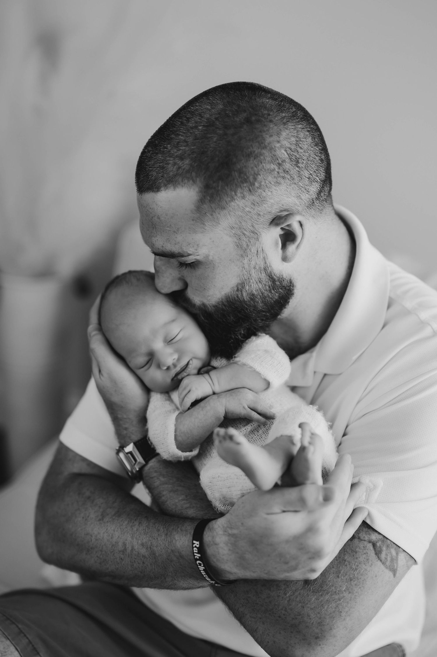 Black and white photo of a bearded father in a polo shirt tenderly kissing the head of his sleeping newborn baby while cradling the infant.
