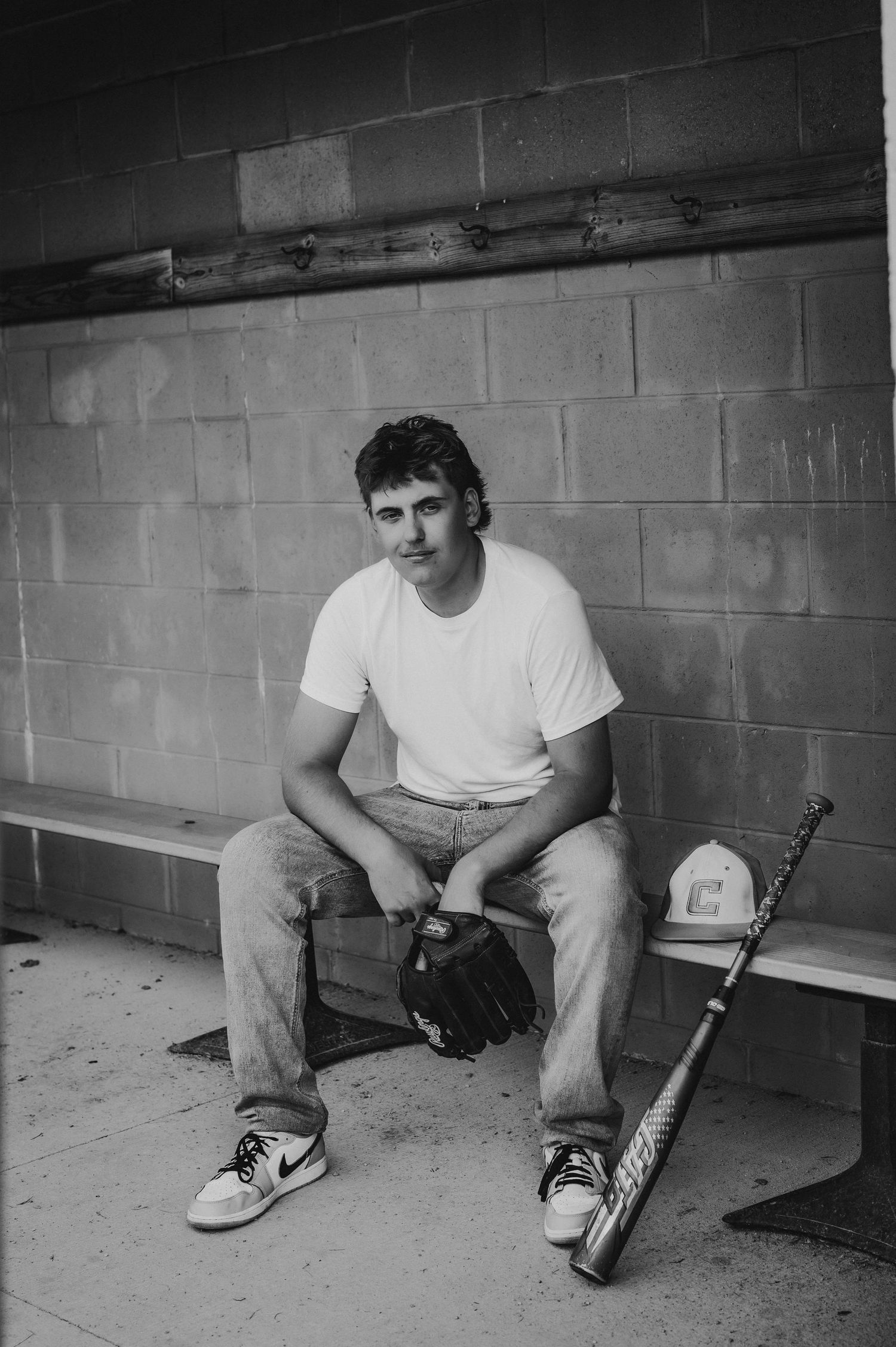 Black and white photo of a young baseball player sitting in a dugout holding a glove with a bat and cap beside him.