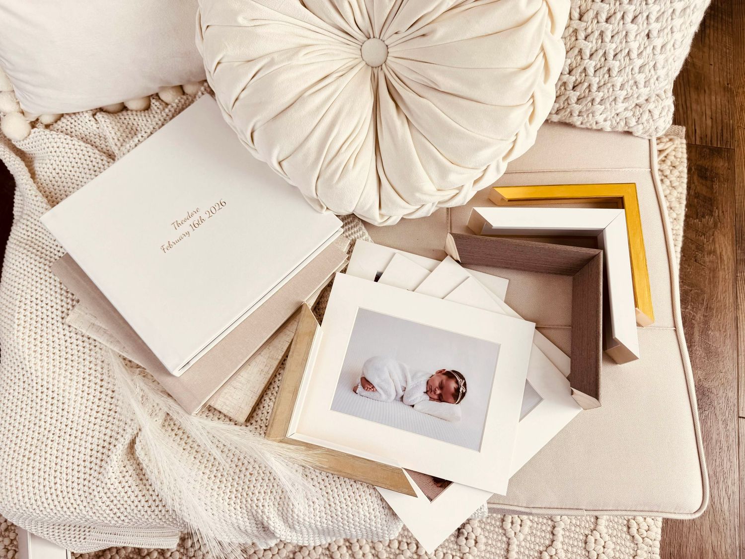 Elegant newborn photo album and framed prints displayed on a cozy beige blanket with a velvet round pillow.