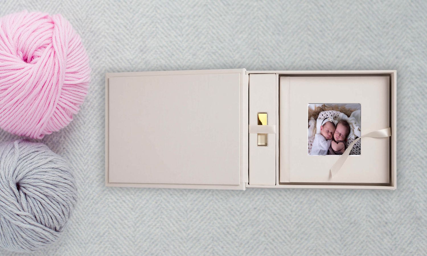 A beige photo keepsake box with USB drive and small print, surrounded by pink and gray yarn balls on a light surface.