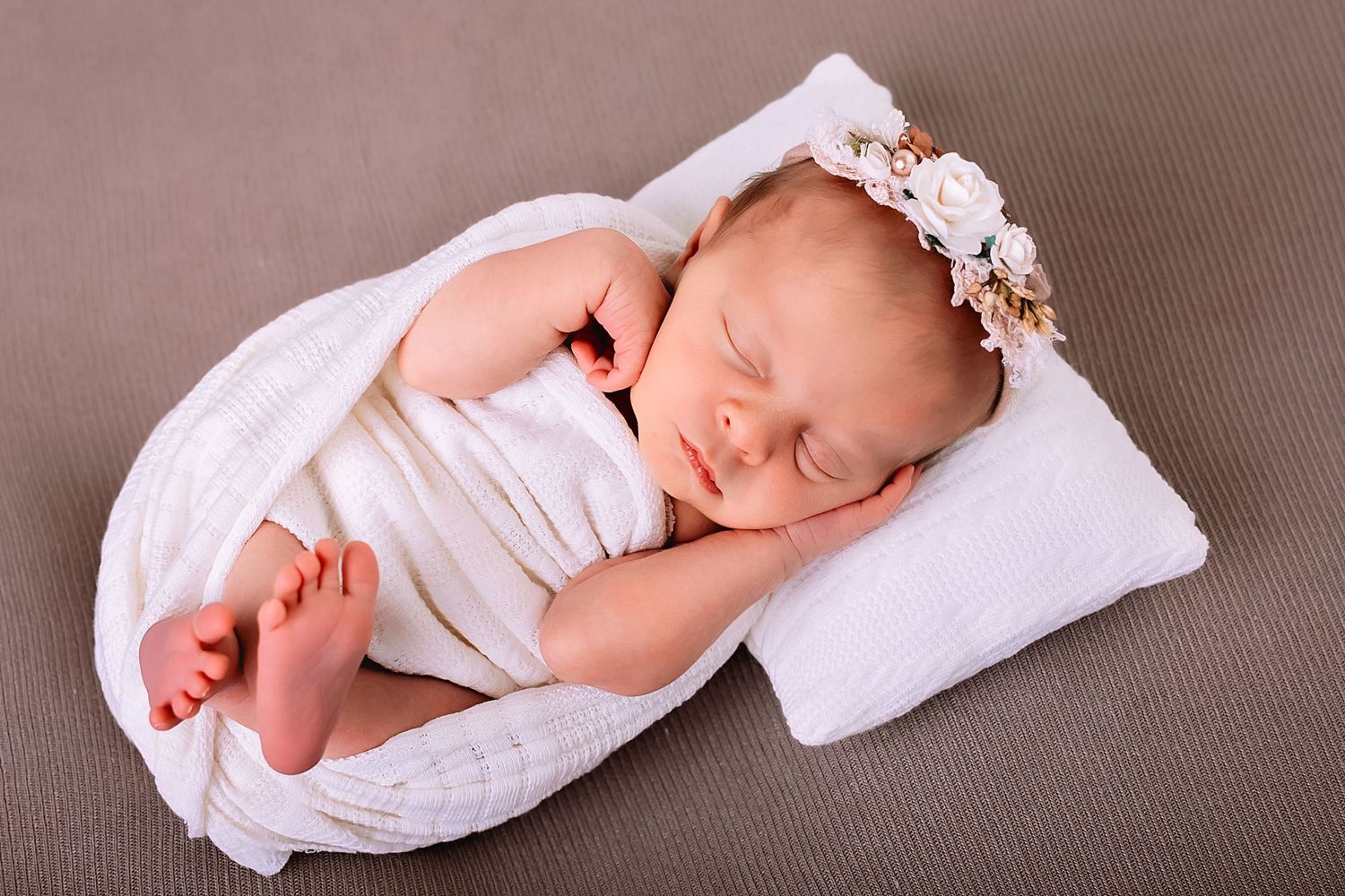 Peaceful newborn baby sleeping wrapped in white knit blanket with floral headband on white pillow.