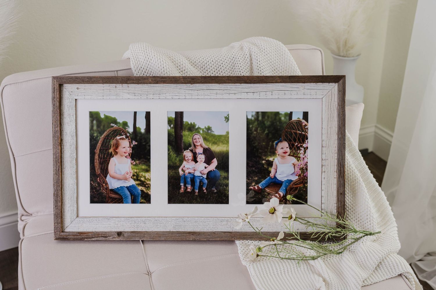 A rustic wooden frame displaying three family portrait photos on a white chair with a soft blanket and wildflowers nearby.