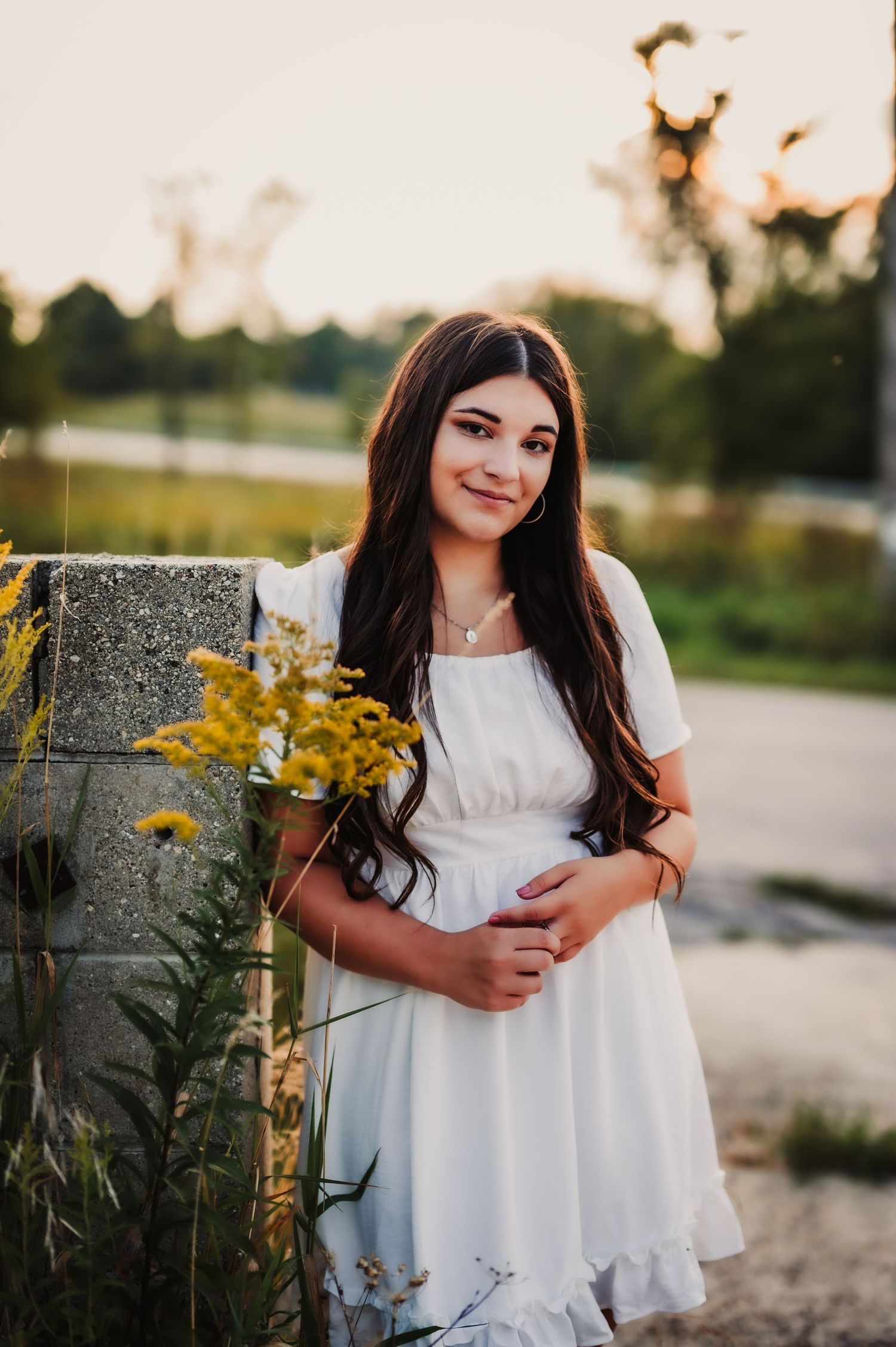 White summer dress contrasts against yellow wildflowers and stone wall at sunset.