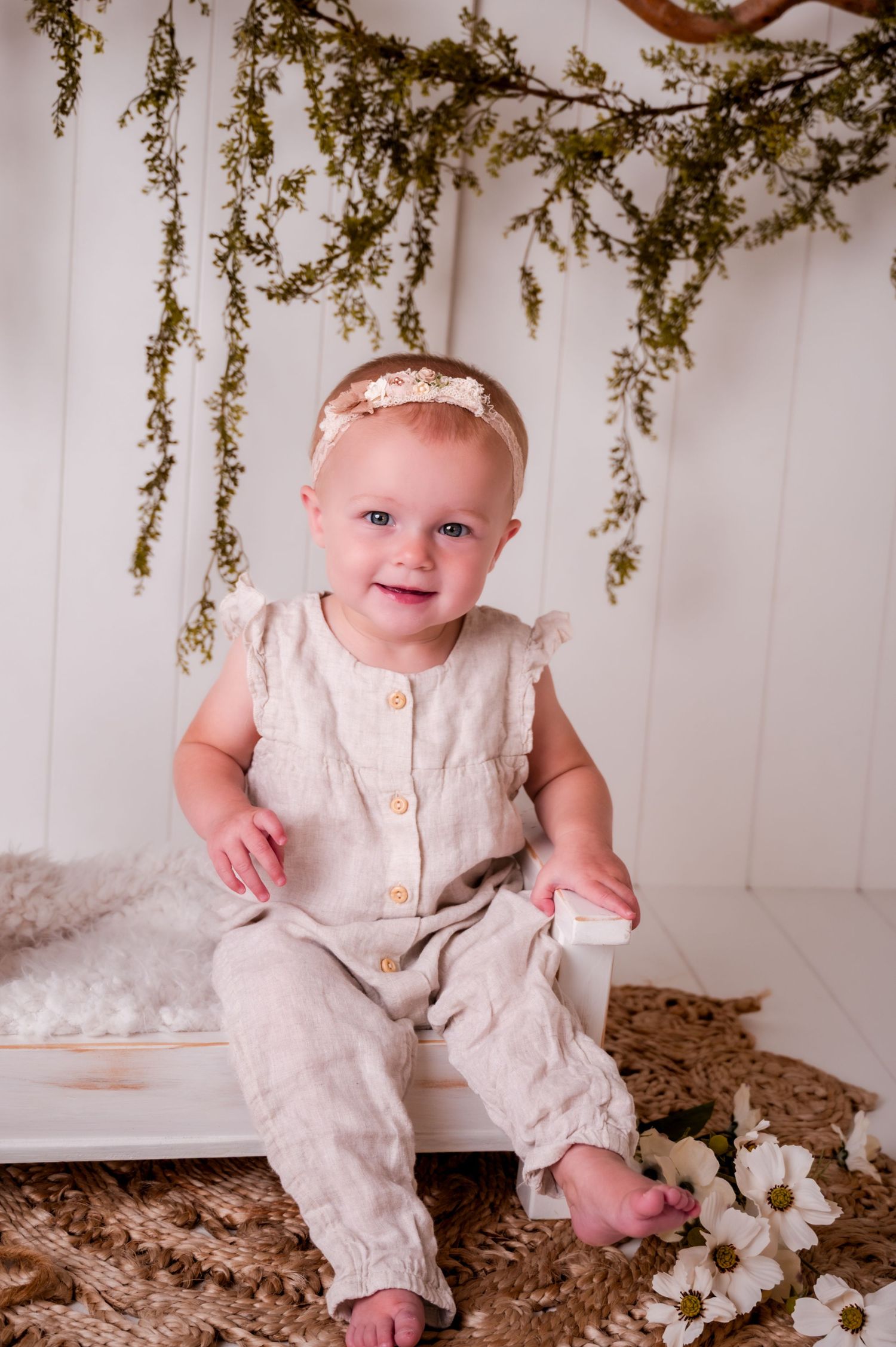 A baby in a cream-colored romper sits on a floor with scattered leaves and white flowers in a Kewaskum, WI  studio setting.