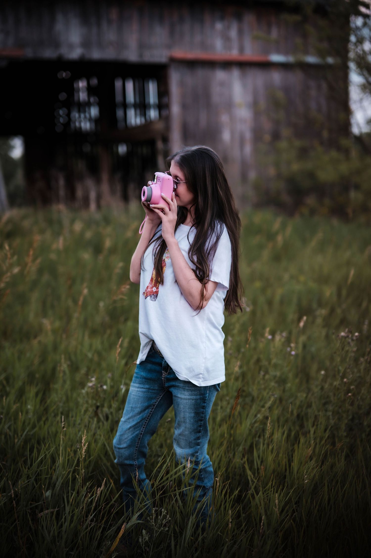 Young woman in white tee and jeans holds pink Instax camera in grassy field near rustic barn at dusk.