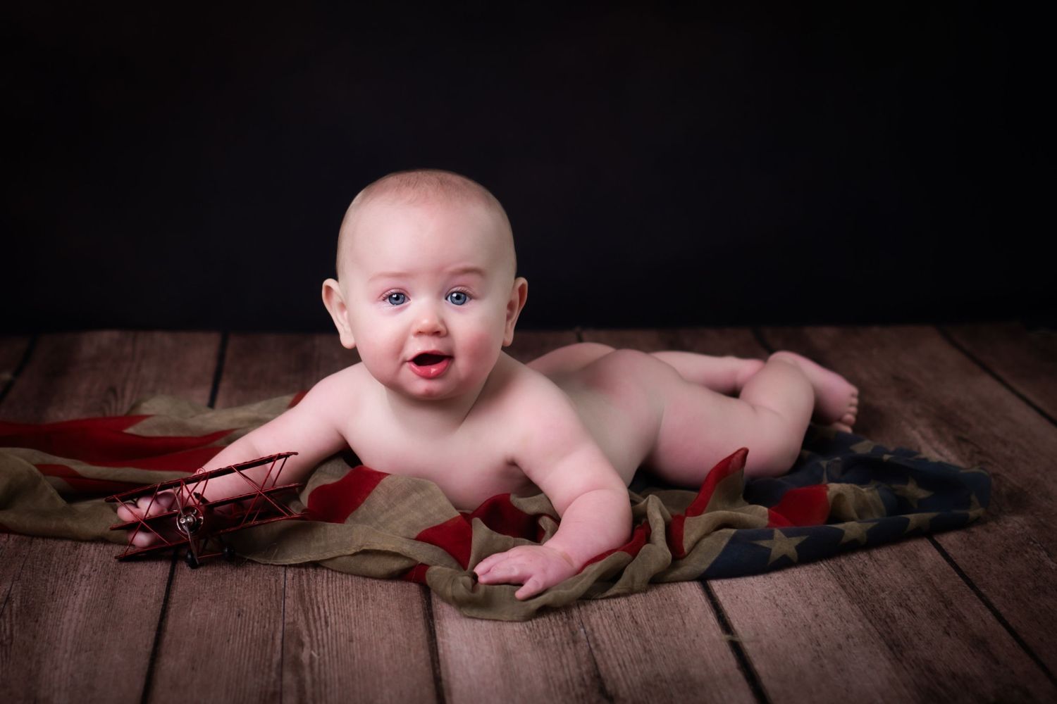 A baby lies on wooden floor surrounded by colorful toy dinosaurs in studio setting.