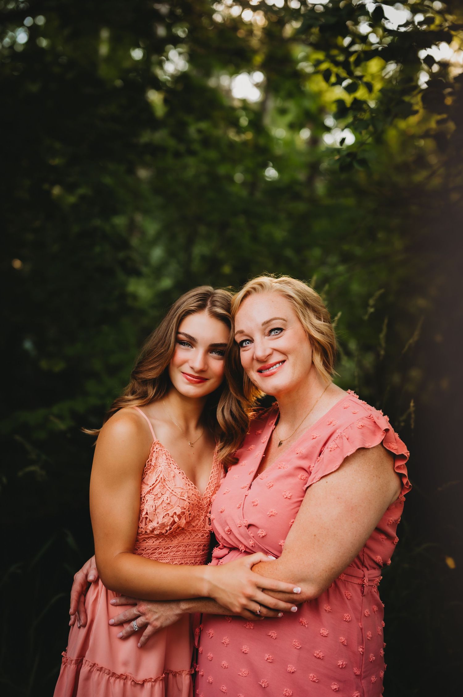 Two people in coordinating pink dresses embrace while posing in natural wooded setting.