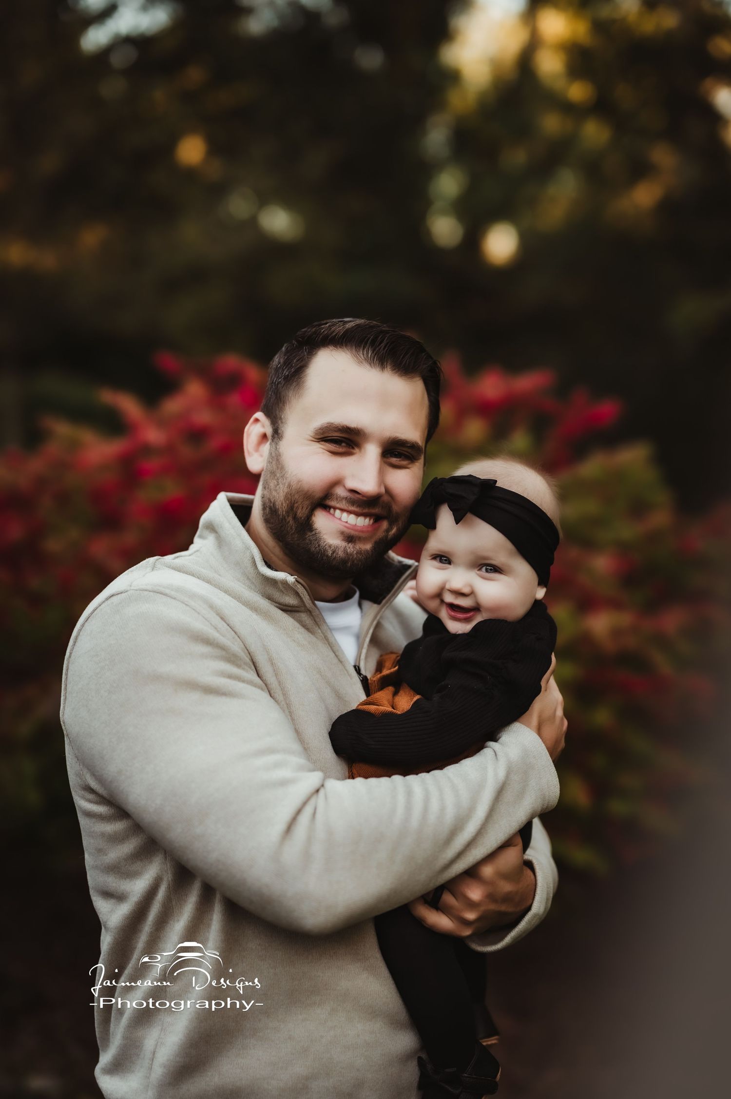 Happy parent and baby share a joyful moment outdoors with red fall foliage in background.