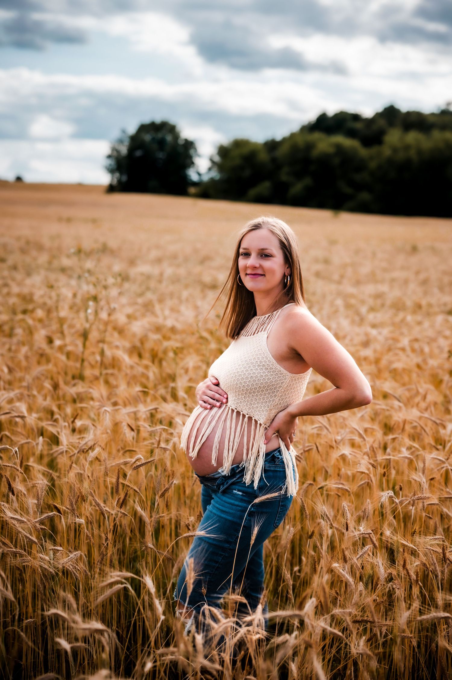 Pregnant woman in crochet top and jeans cradling baby bump in golden wheat field under cloudy sky.