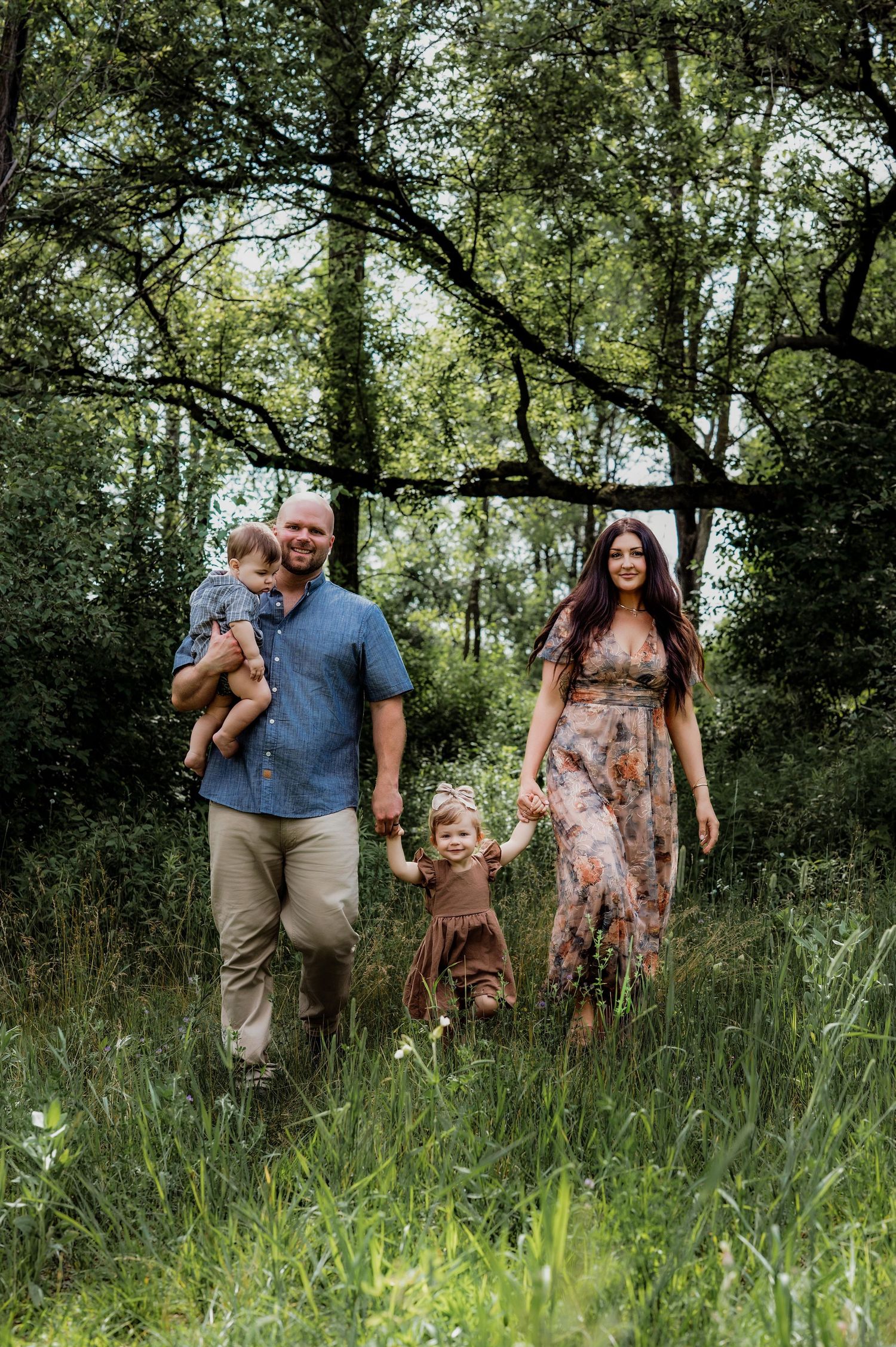 A smiling family of four walks through a lush green forest, parents holding children's hands on a sunny day.
