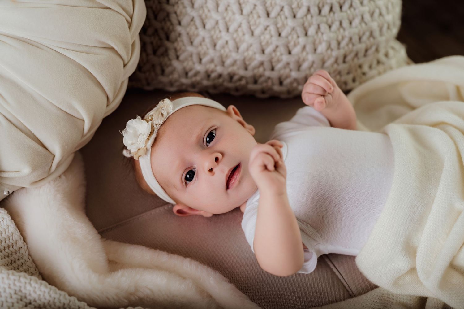 Adorable baby girl in white onesie and floral headband lying on a cozy beige blanket looking up curiously.