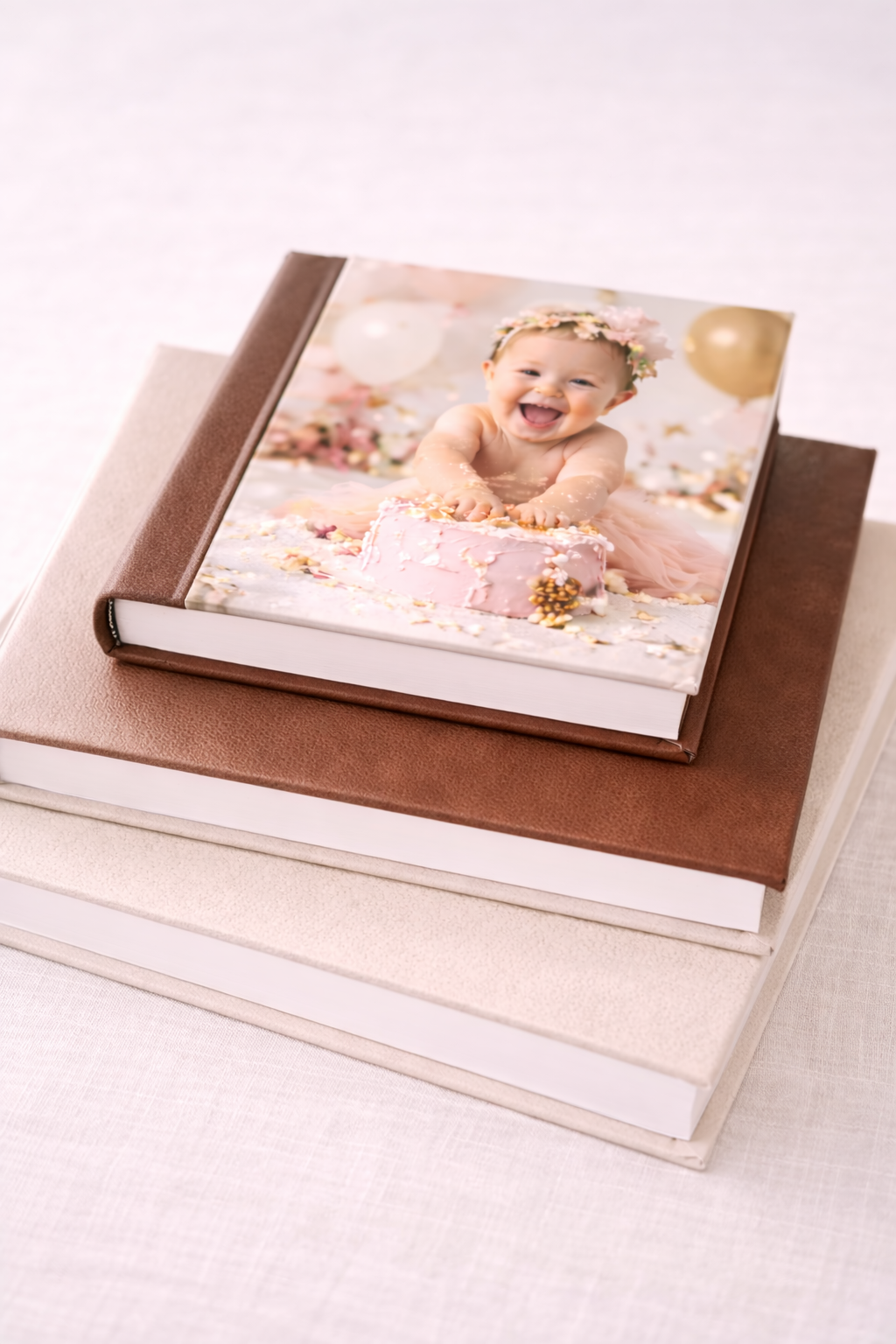 Stack of photo books with top book featuring a smiling baby in floral crown celebrating first birthday with pink cake.