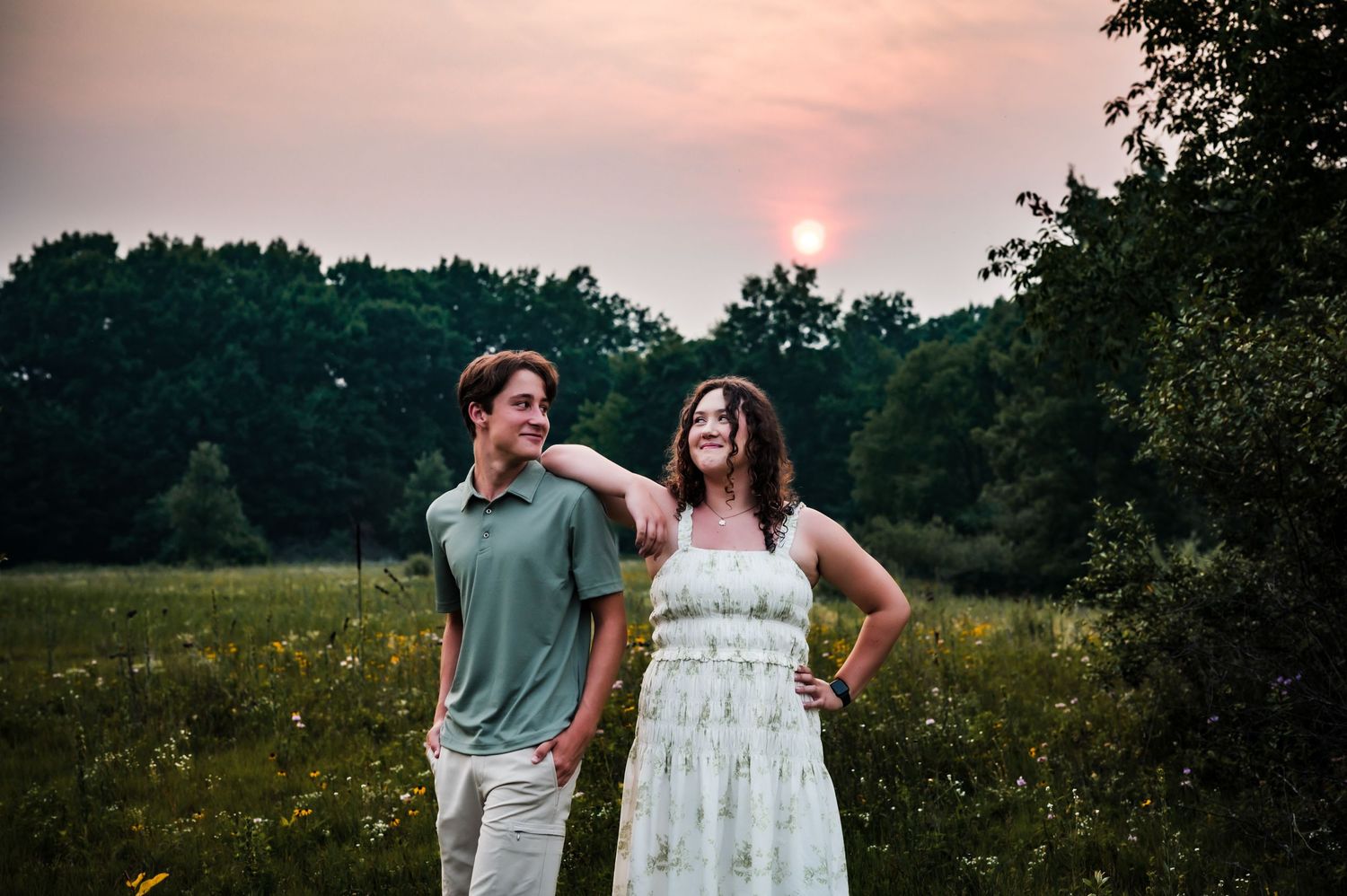 Couple smiling at sunset in a wildflower meadow, surrounded by lush green trees in warm golden light.