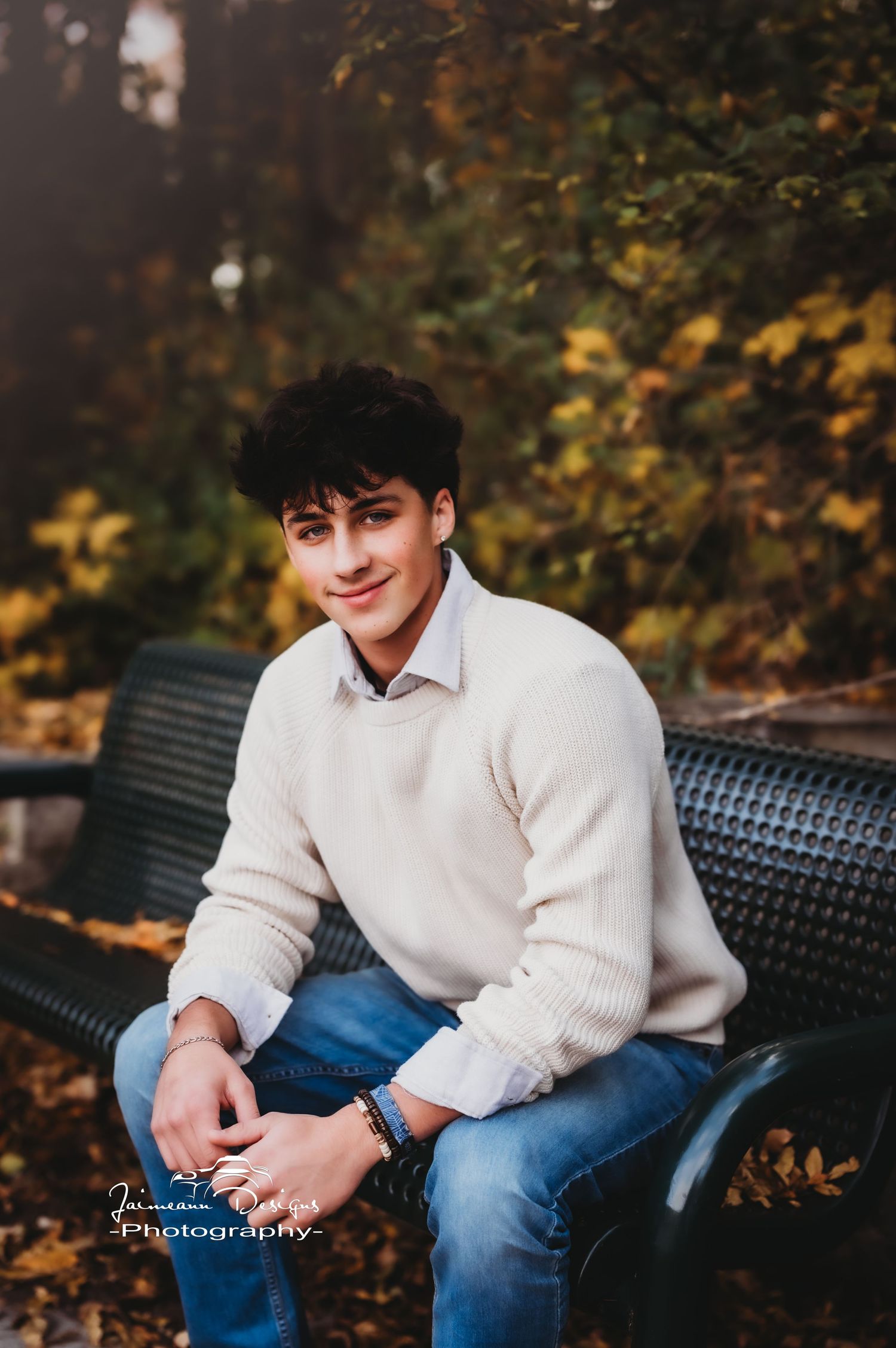 Seated portrait on black bench wearing cream sweater and jeans in autumn setting.