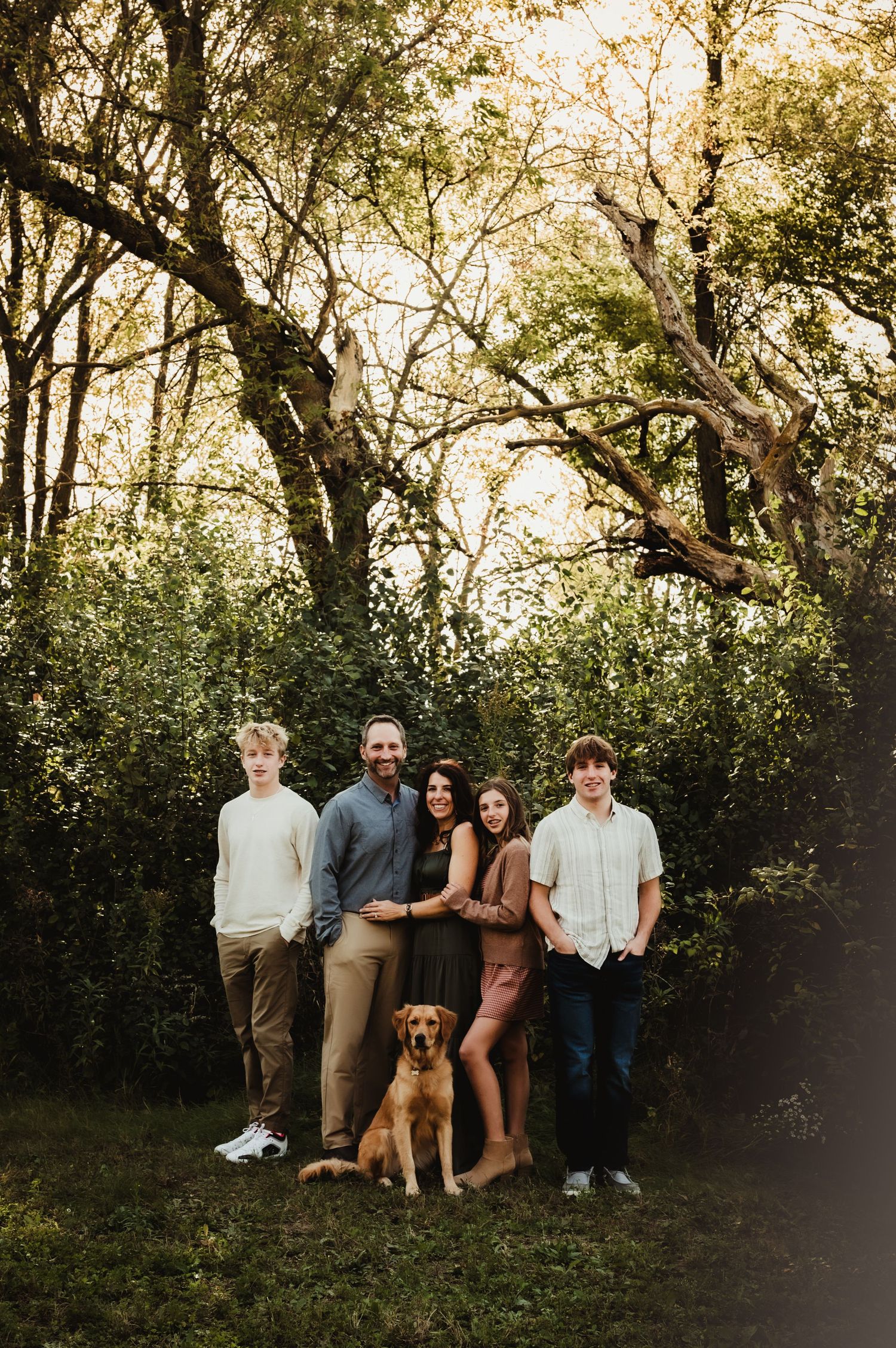 Family of four with a golden retriever dog posing outdoors among lush green trees during golden hour.