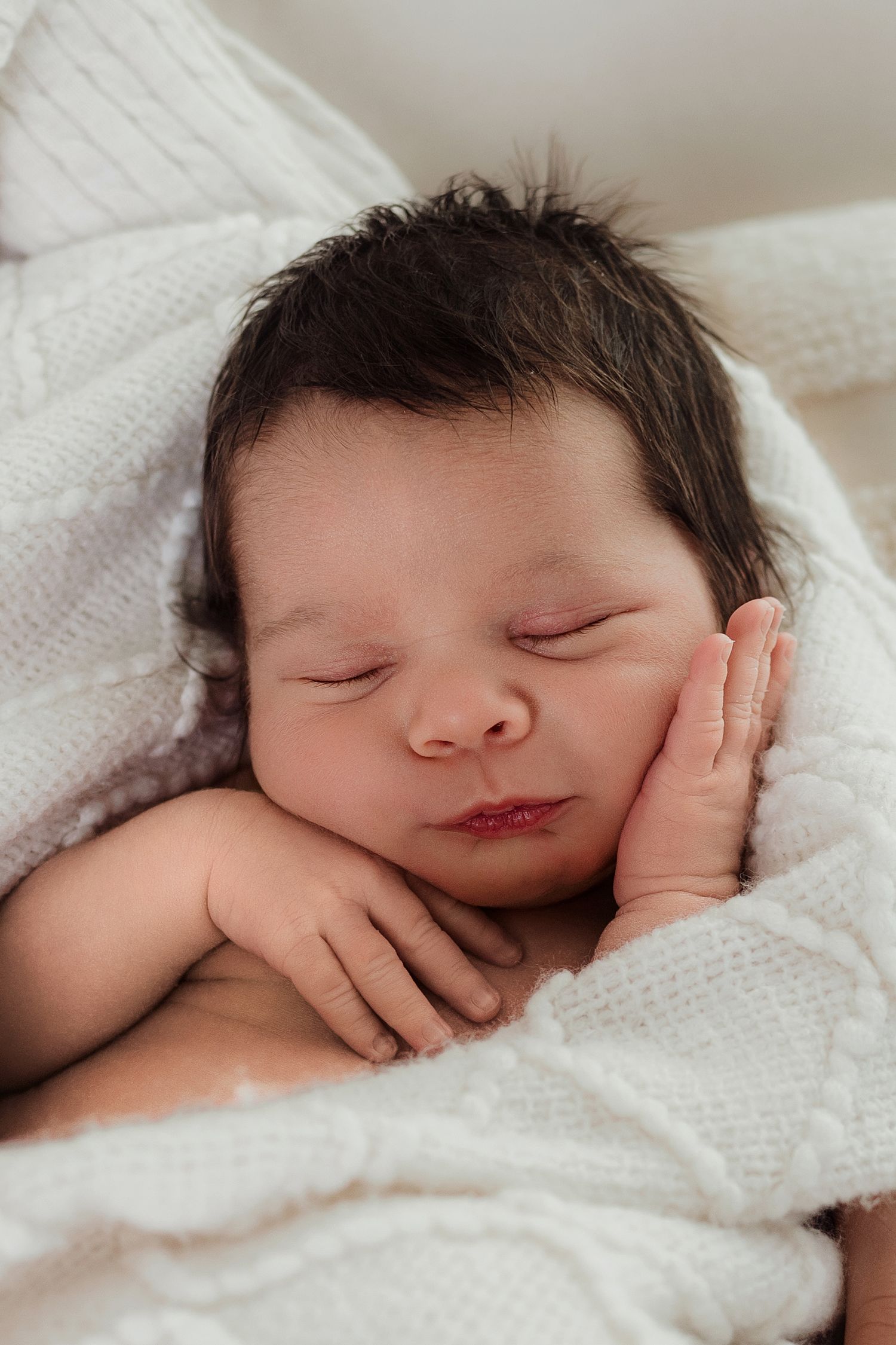 Peaceful newborn baby with dark hair sleeping on a white knit blanket with tiny hands tucked under cheek.