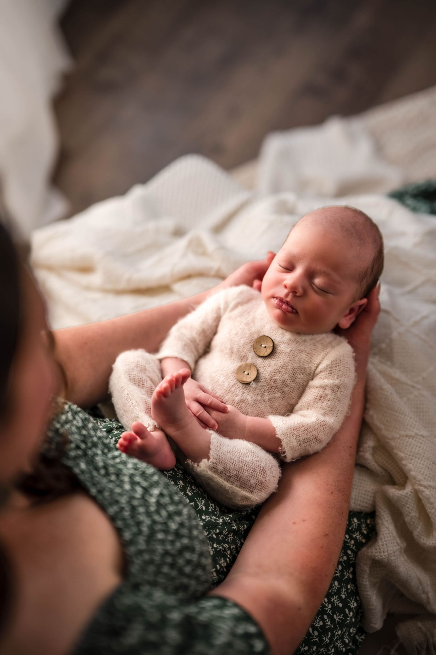 Sleeping newborn baby in a cream knit romper with wooden buttons, cradled in a mother's hands on a soft white blanket.
