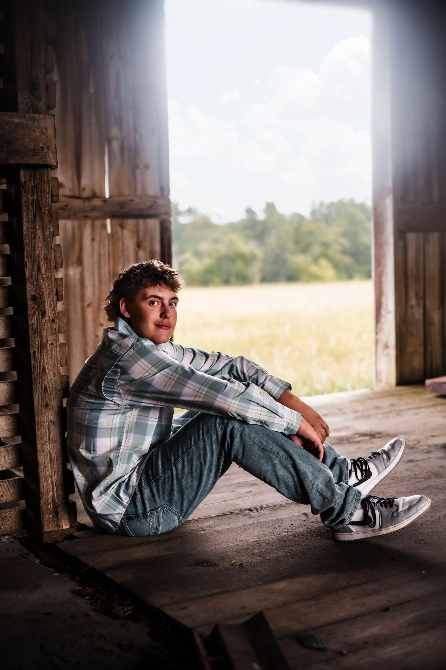 Young man in plaid shirt and jeans sitting in rustic barn doorway with scenic field view behind him.