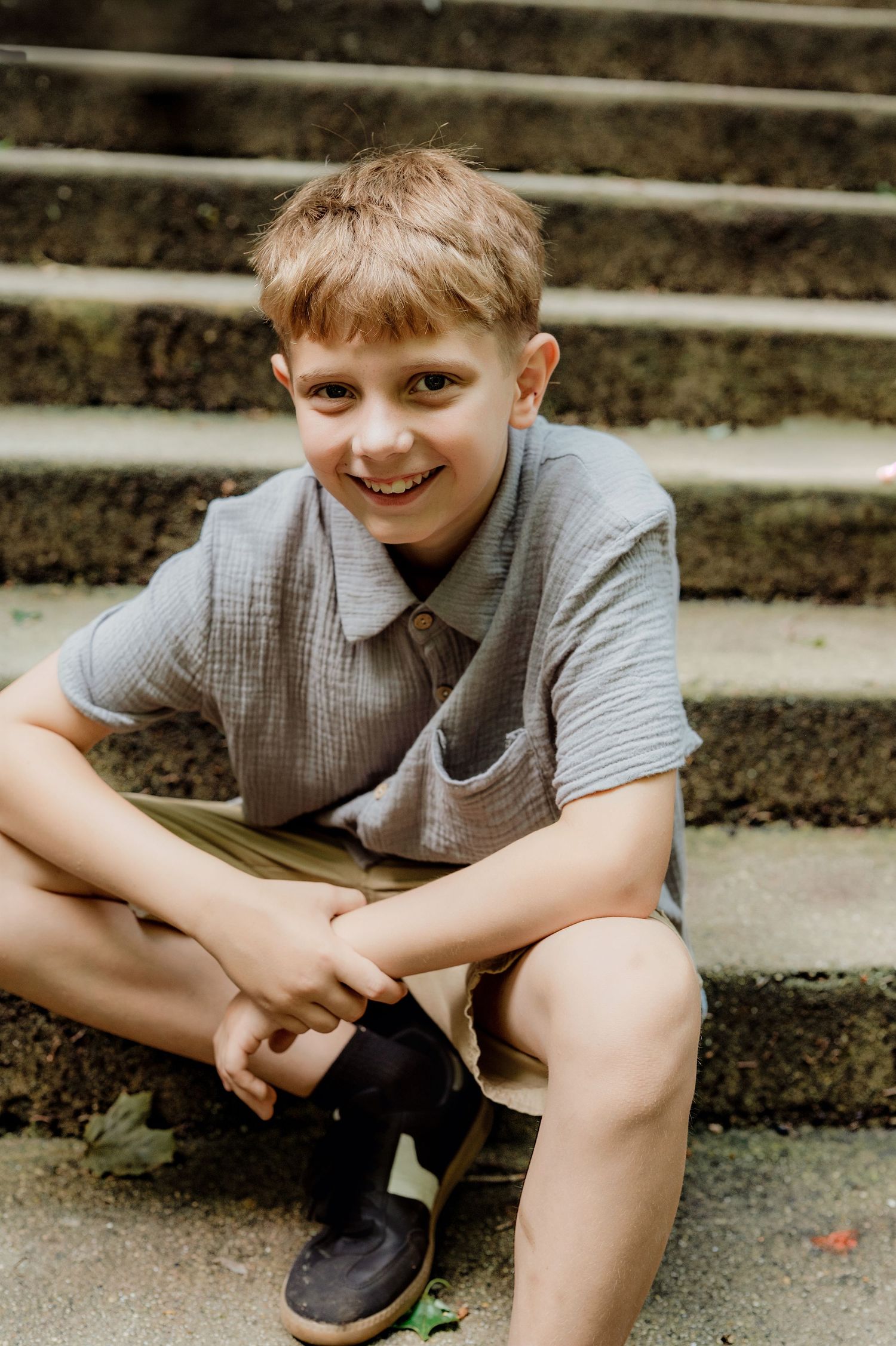 Smiling boy sitting on stone steps outdoors, wearing a gray shirt and shorts, posing casually for a portrait photo.