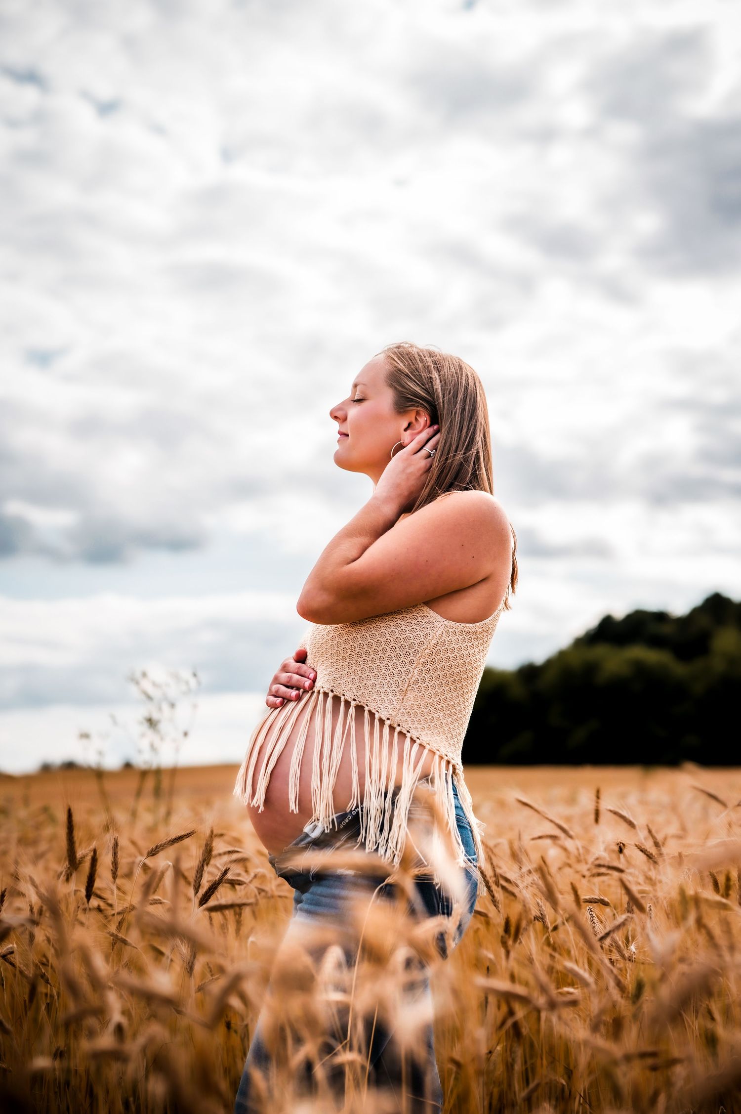 Pregnant woman standing in golden wheat field wearing crochet top, eyes closed, hand on neck, cloudy sky background.
