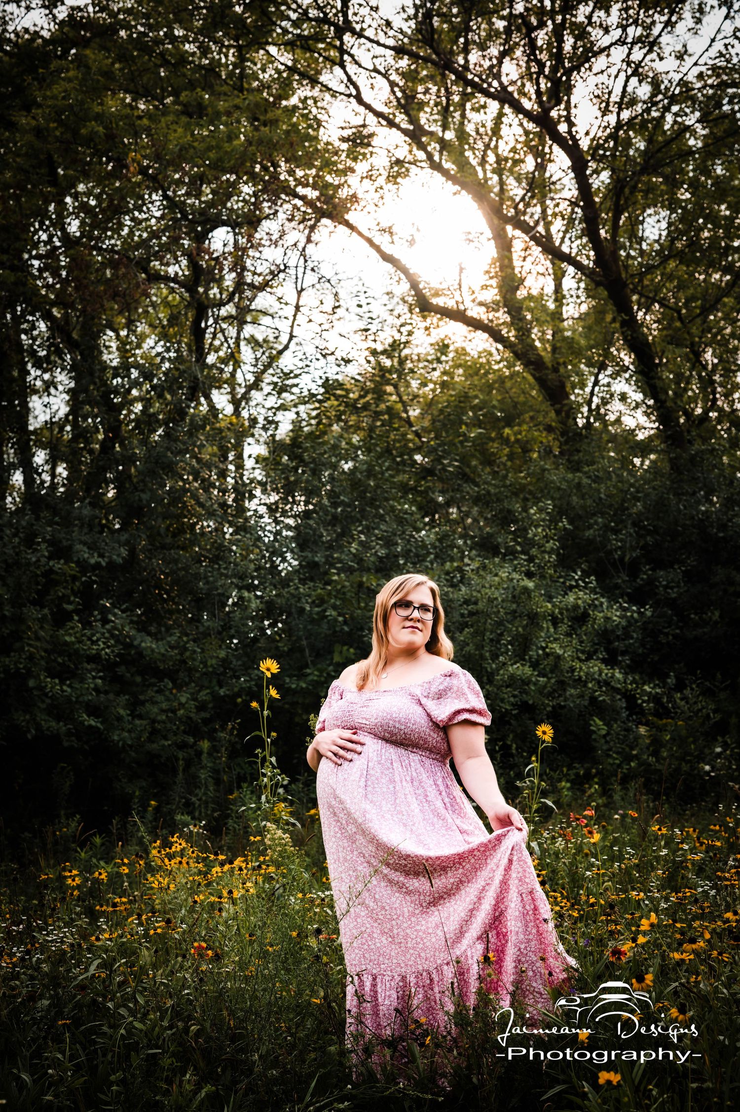 Pregnant woman in pink floral maxi dress standing in wildflower field surrounded by lush green trees at golden hour.