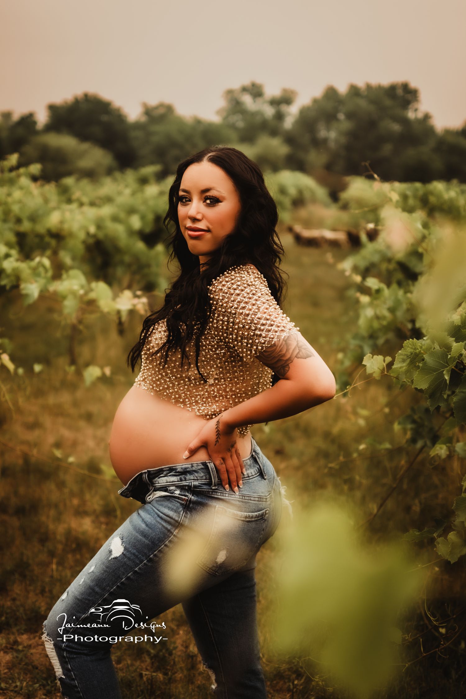 Maternity photo of an expectant mother posing in a vineyard wearing a sequined top and distressed jeans at sunset.