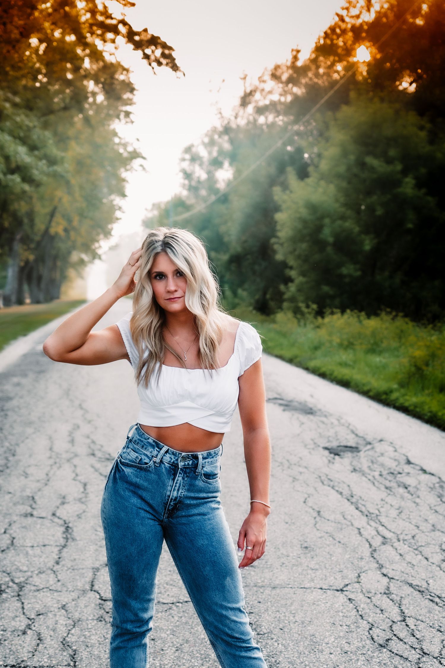 Person in white crop top and jeans poses on cracked rural road at sunset.