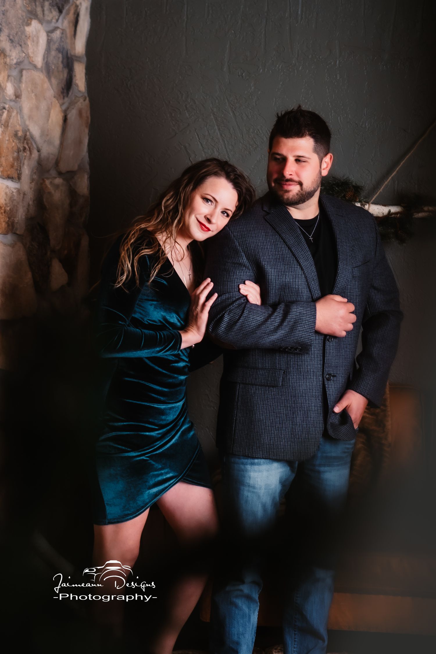 Romantic couple pose against stone wall, woman in velvet dress and man in gray blazer.