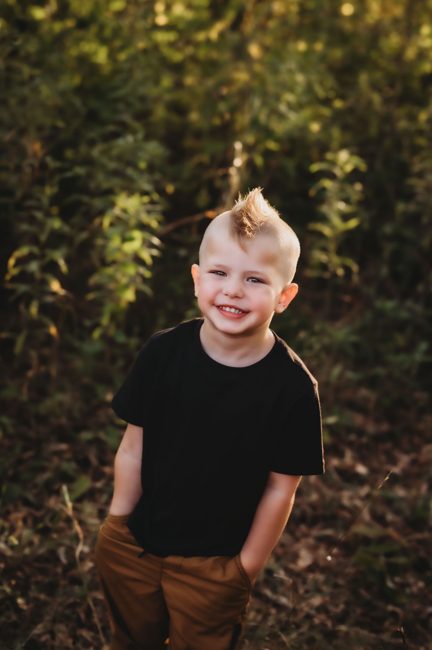 Young child in black t-shirt and brown pants smiles while standing in front of greenery in outdoor portrait setting.