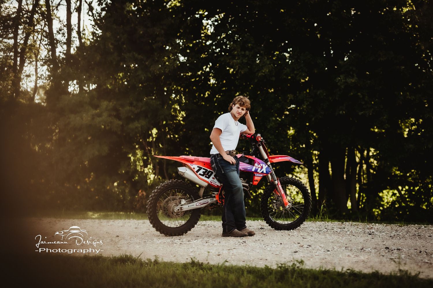 A dirt bike stands in a sunlit forest clearing on a gravel path.