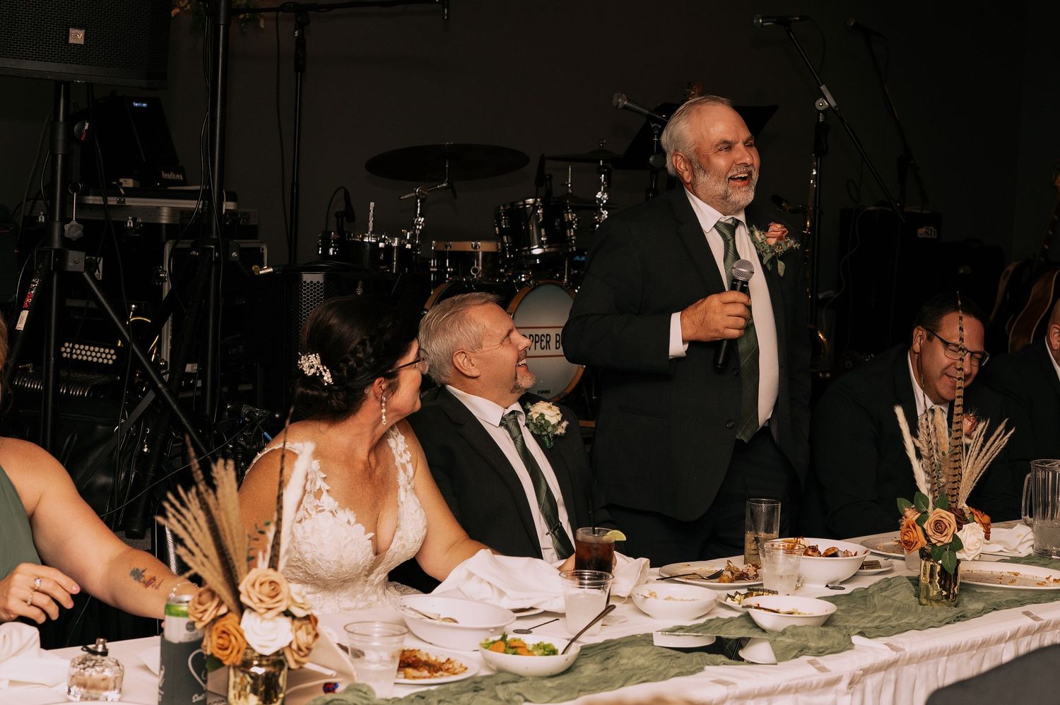 Wedding reception guests enjoy dinner and conversation at a dimly lit banquet table with floral centerpieces.
