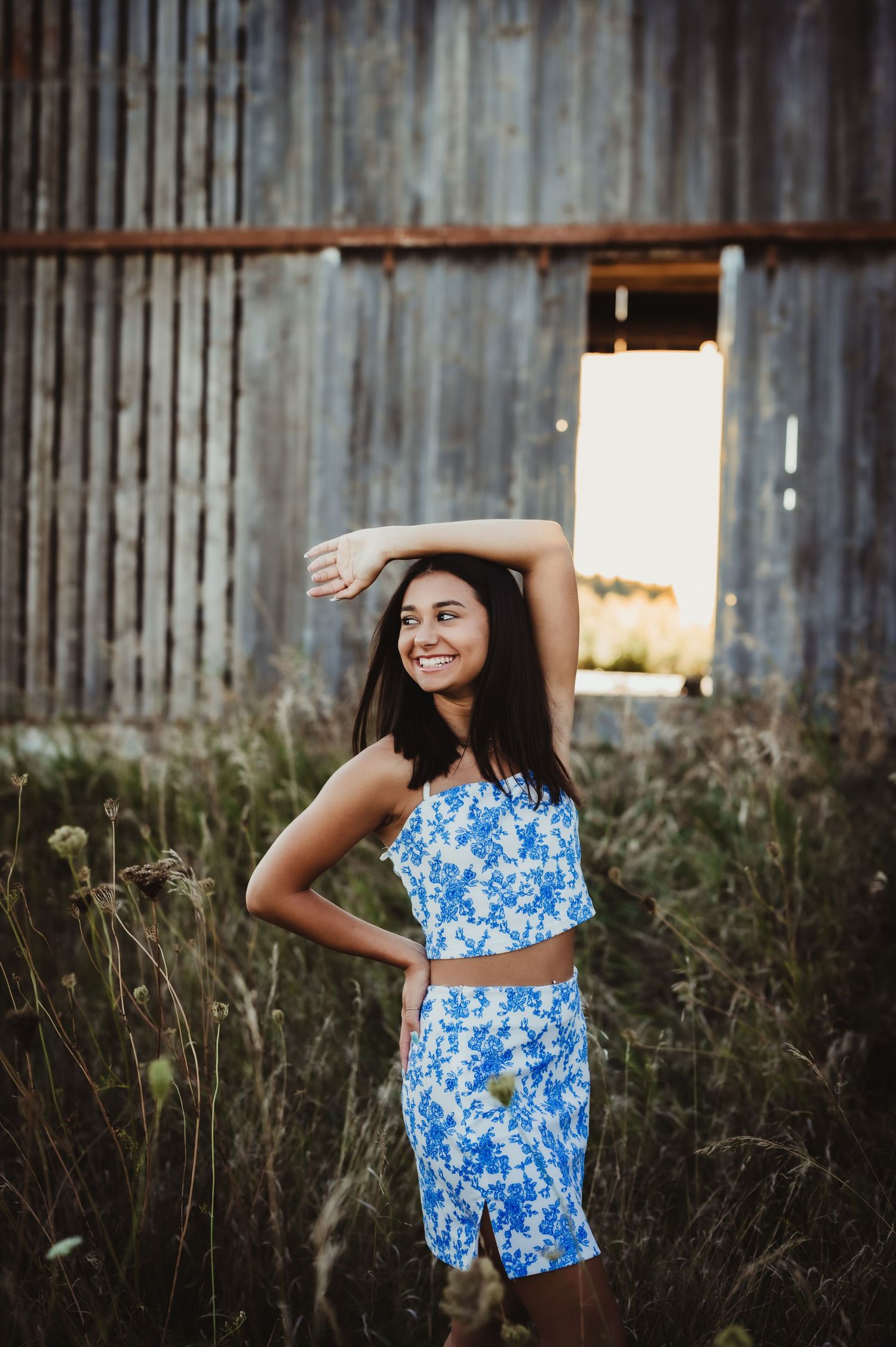 Model in blue two-piece outfit poses playfully by weathered barn wall.