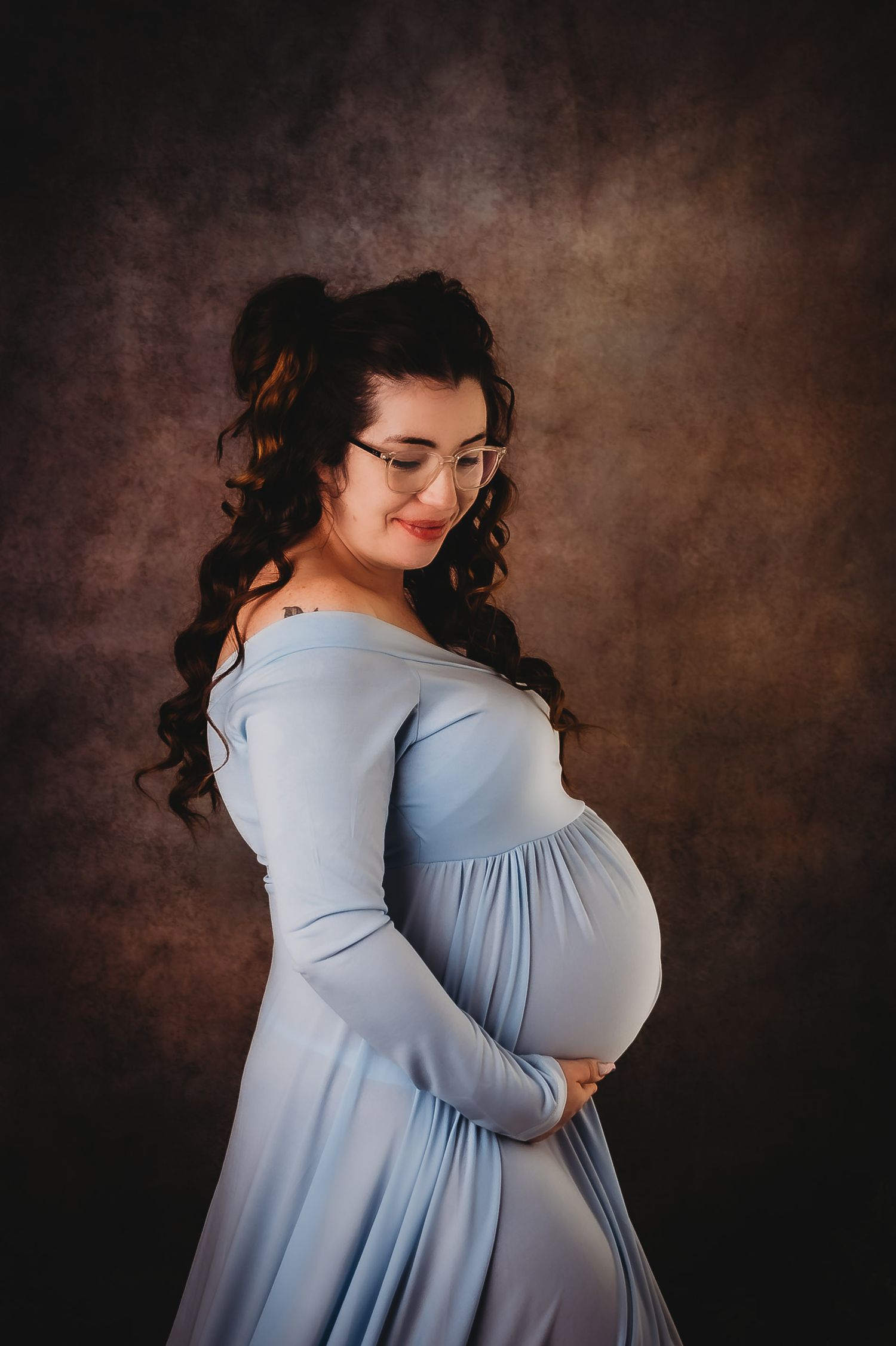 Side profile maternity portrait featuring a powder blue off-shoulder gown against a textured brown background.