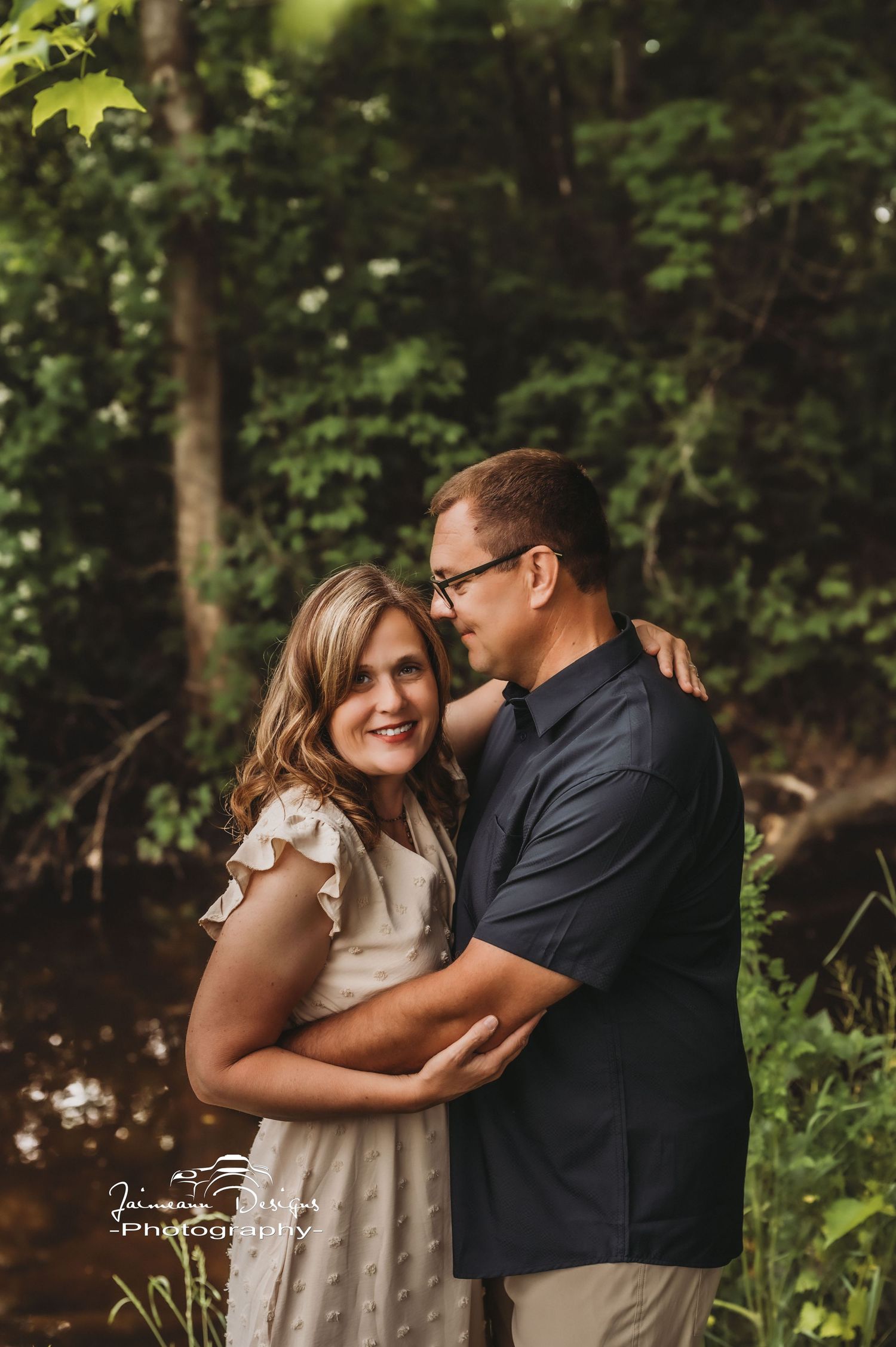 A romantic couple embrace in a wooded setting with soft natural lighting.