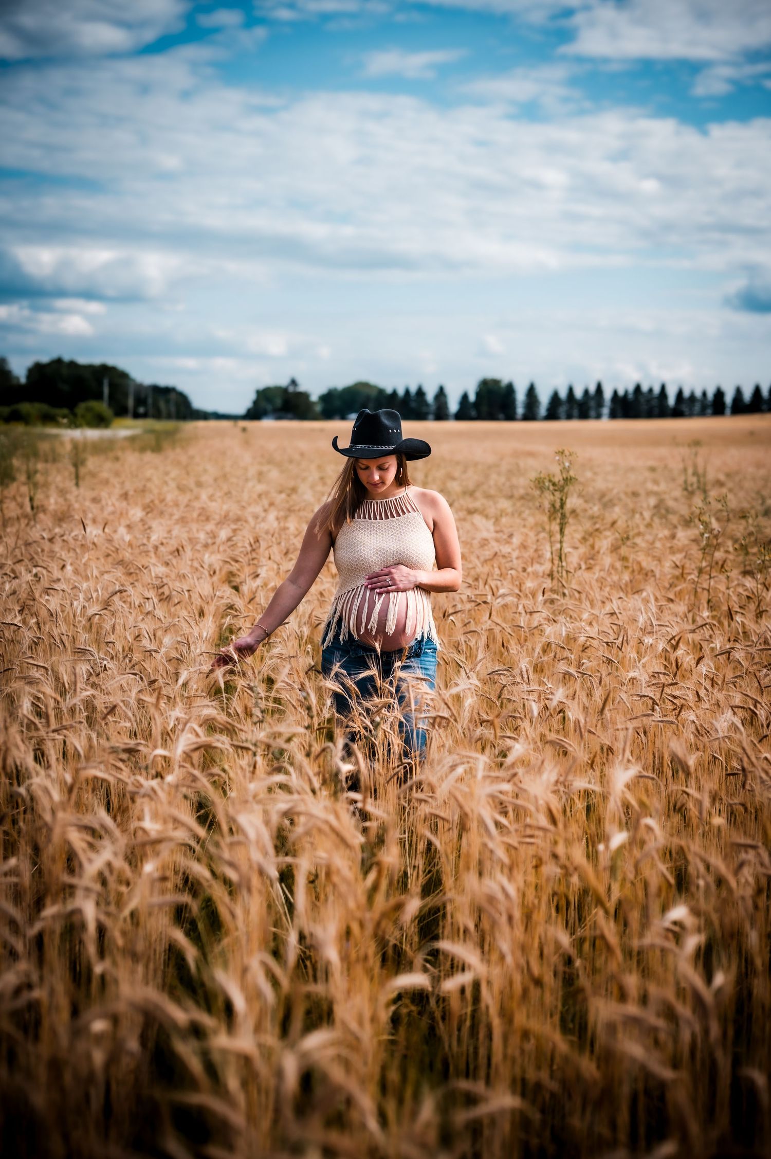 Woman in black cowboy hat walking through golden wheat field under cloudy blue sky.