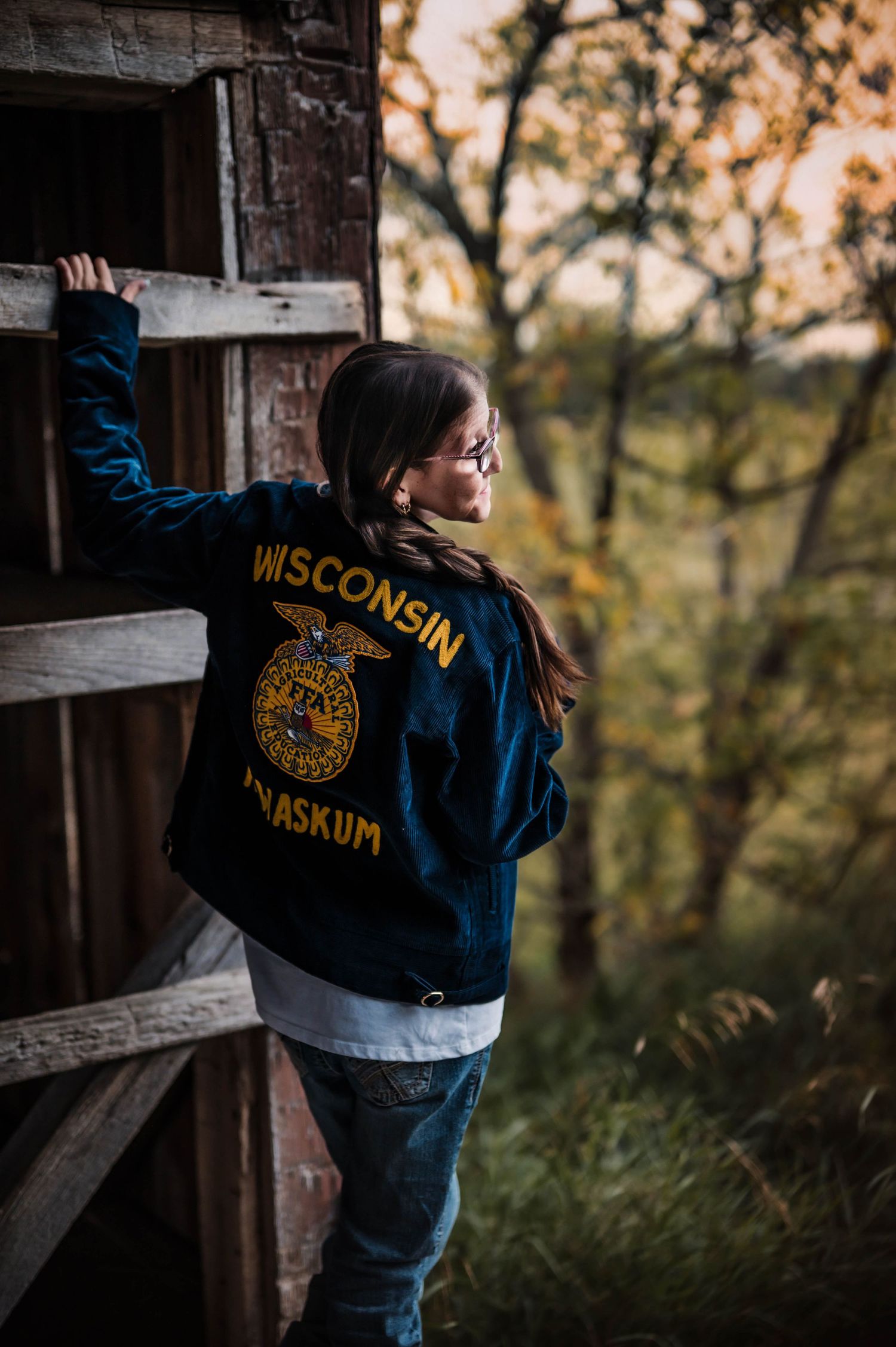 Young woman in Wisconsin FFA jacket leans against rustic barn door surrounded by autumn foliage.