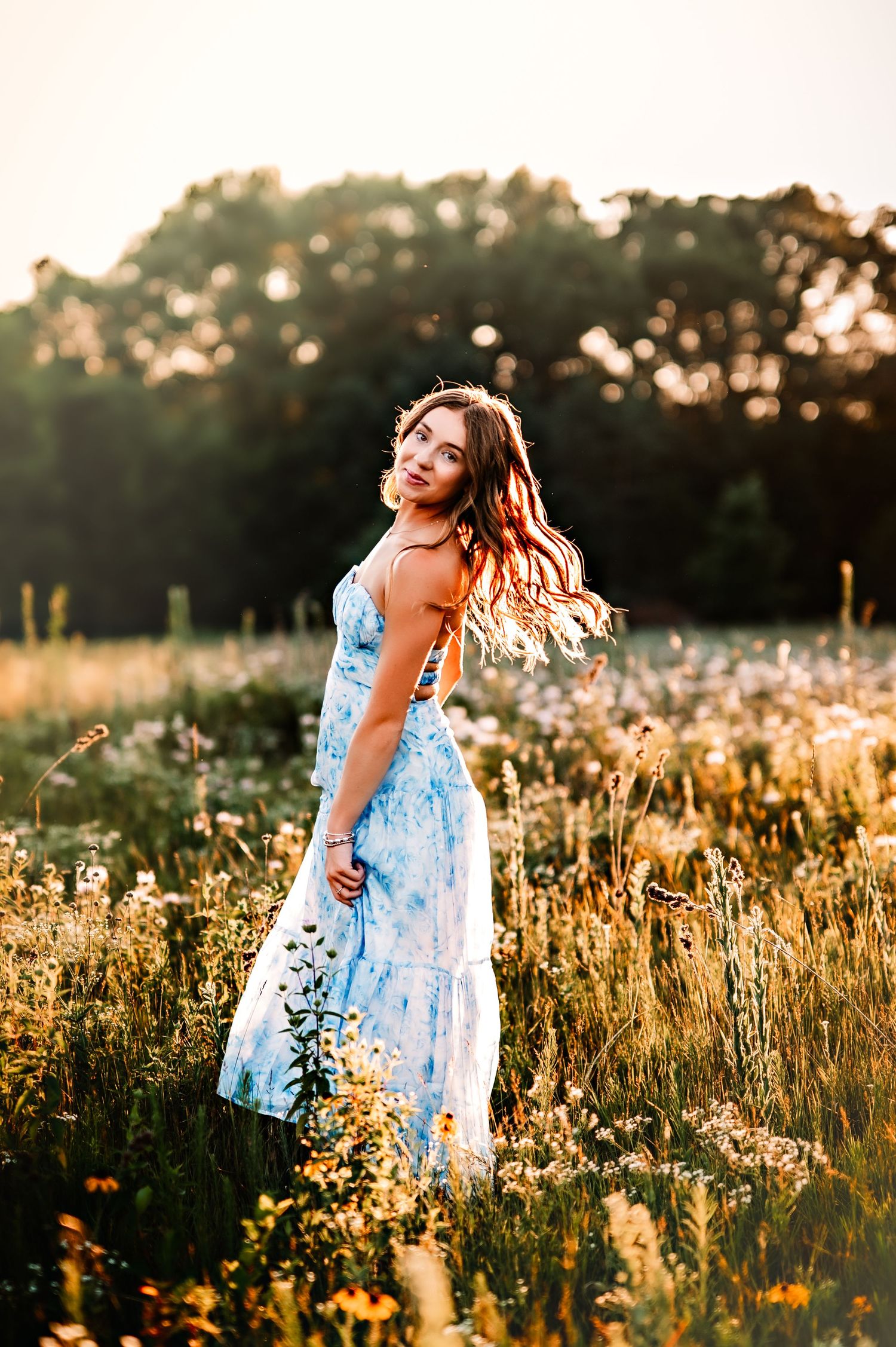 Young woman in blue floral sundress standing in wildflower field at golden hour with trees in background.