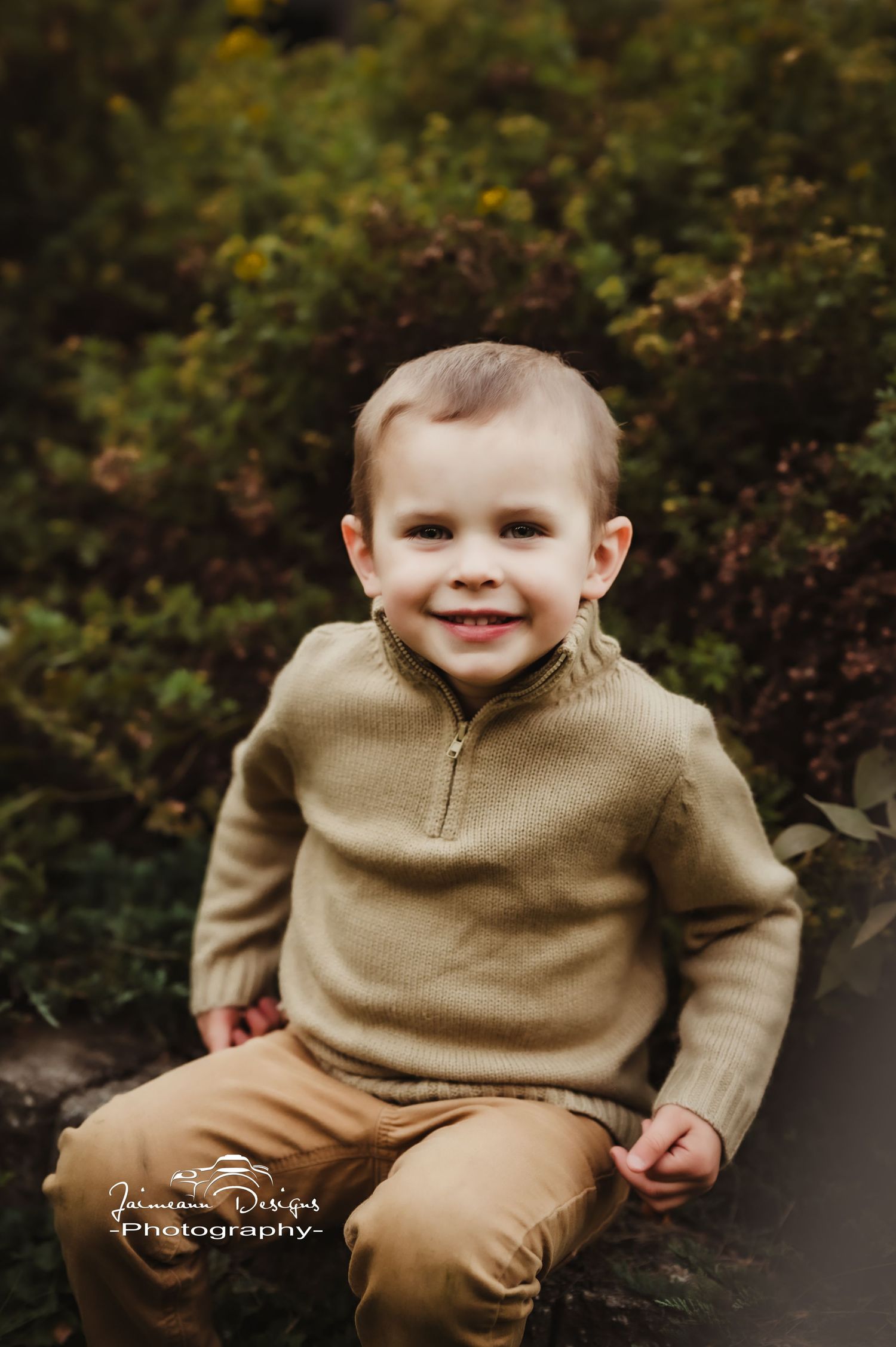 Young child in beige sweater sits on stone step with autumn foliage in background.