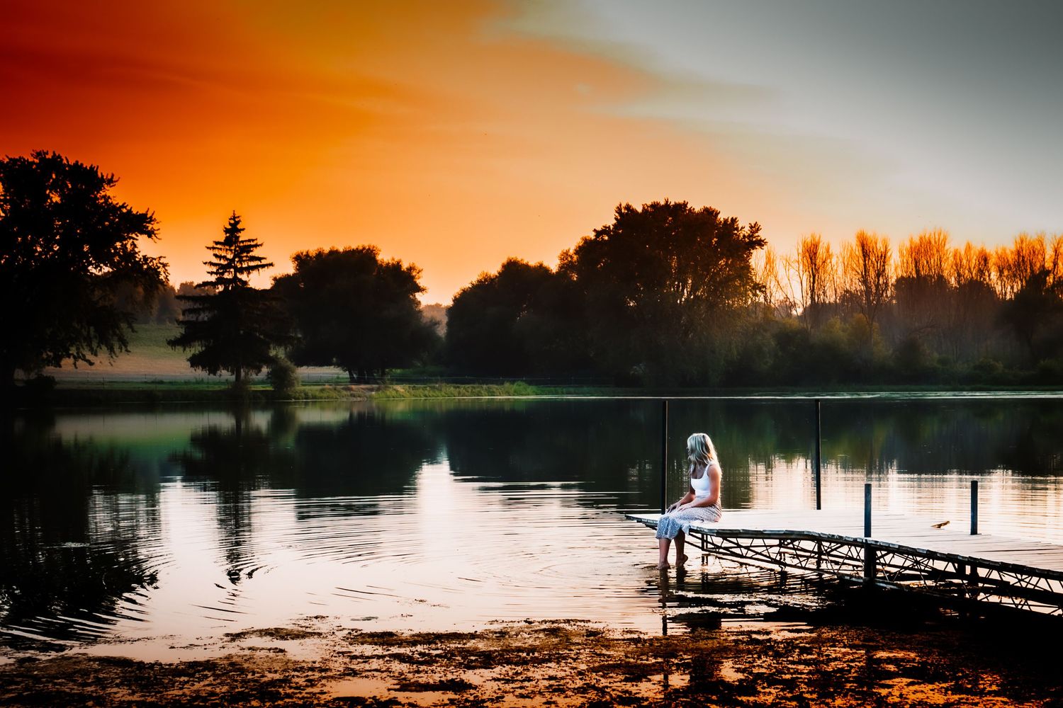 Silhouette on dock at sunset with orange sky reflecting on calm lake.