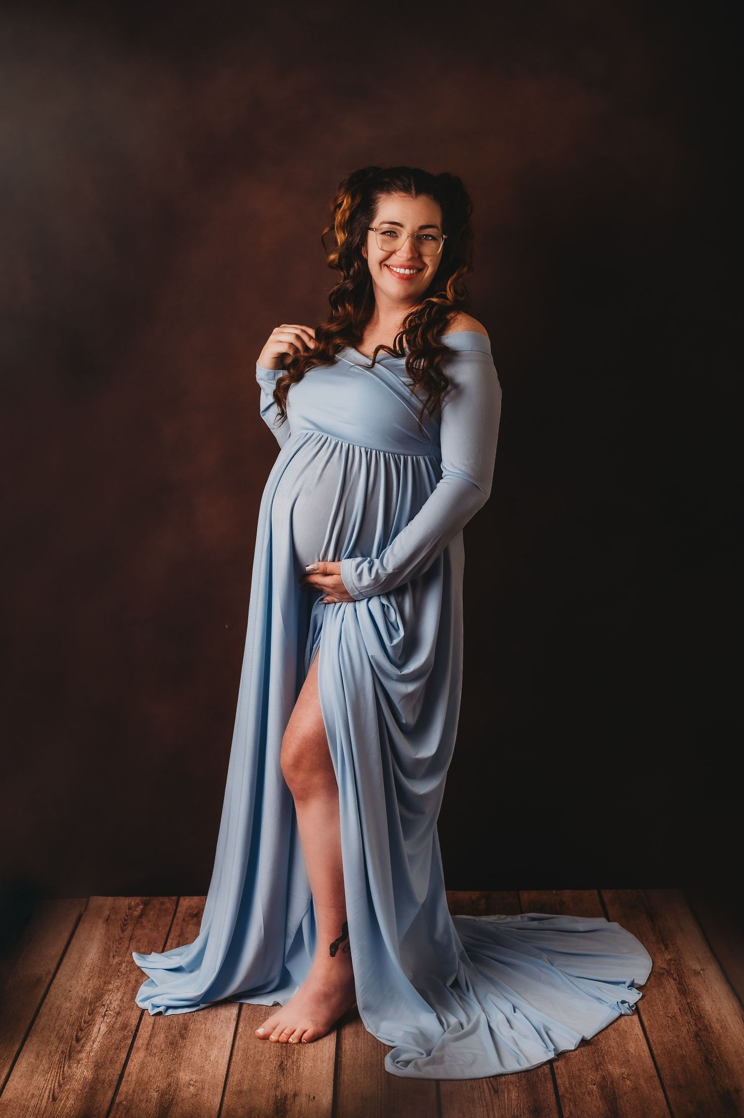 Maternity portrait featuring flowing blue dress with side slit on wooden floor.