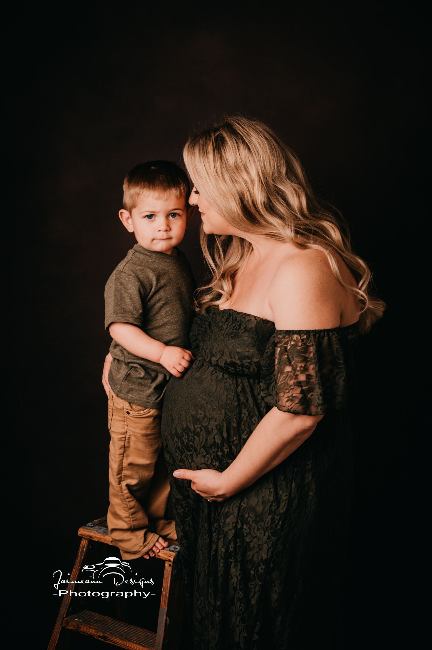 Tender maternity moment captured against dark backdrop with vintage wooden step stool.
