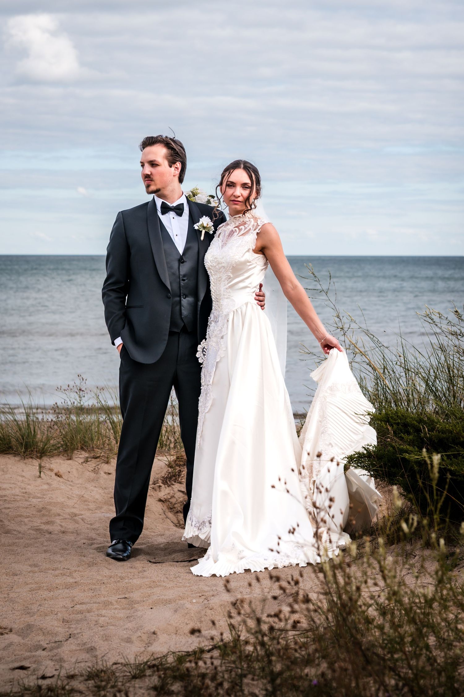 Bride in white lace gown and groom in black tuxedo pose together on sandy beach with ocean backdrop.