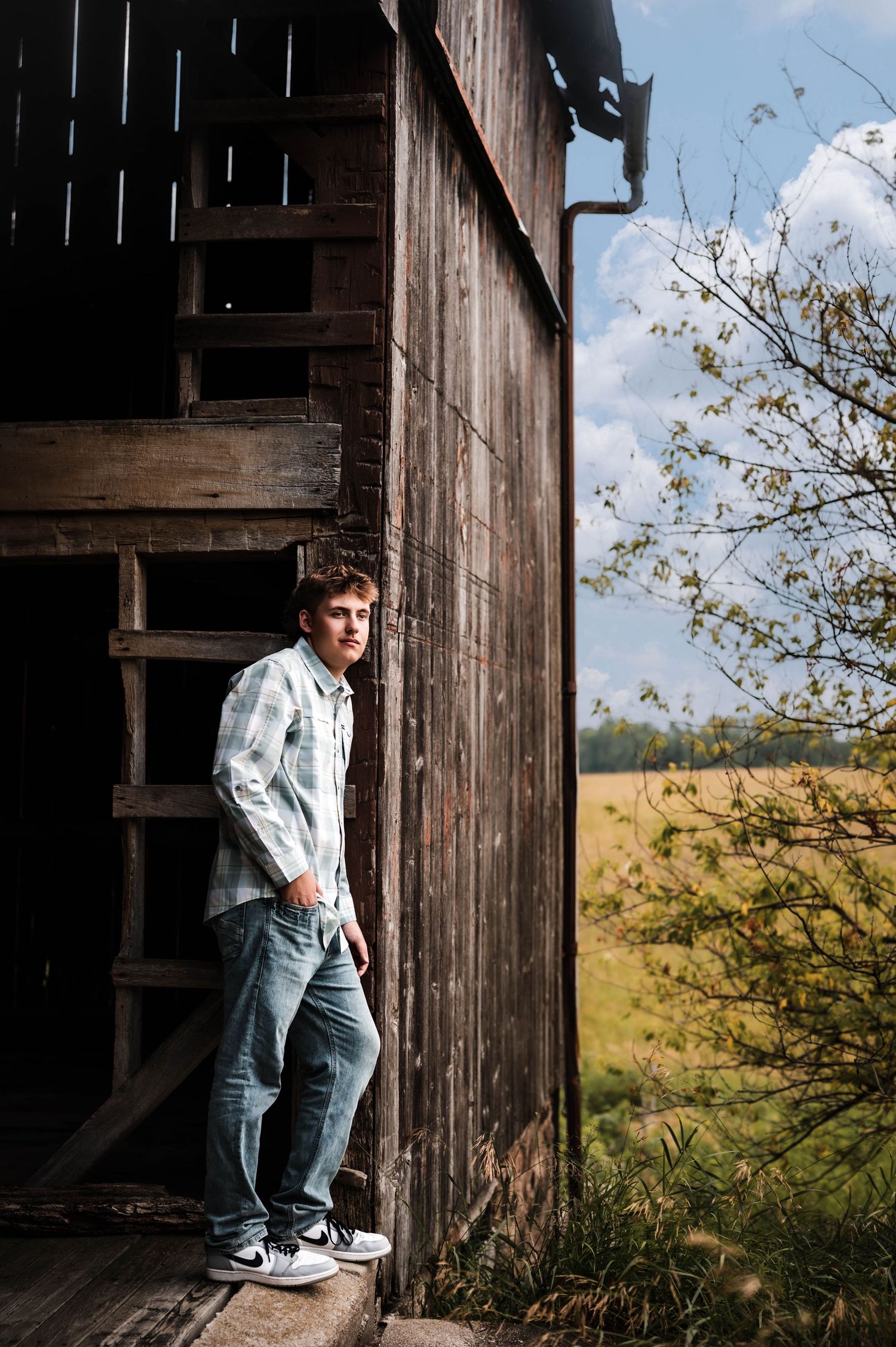 Young man in plaid shirt and jeans leaning against rustic wooden barn with green fields and blue sky.
