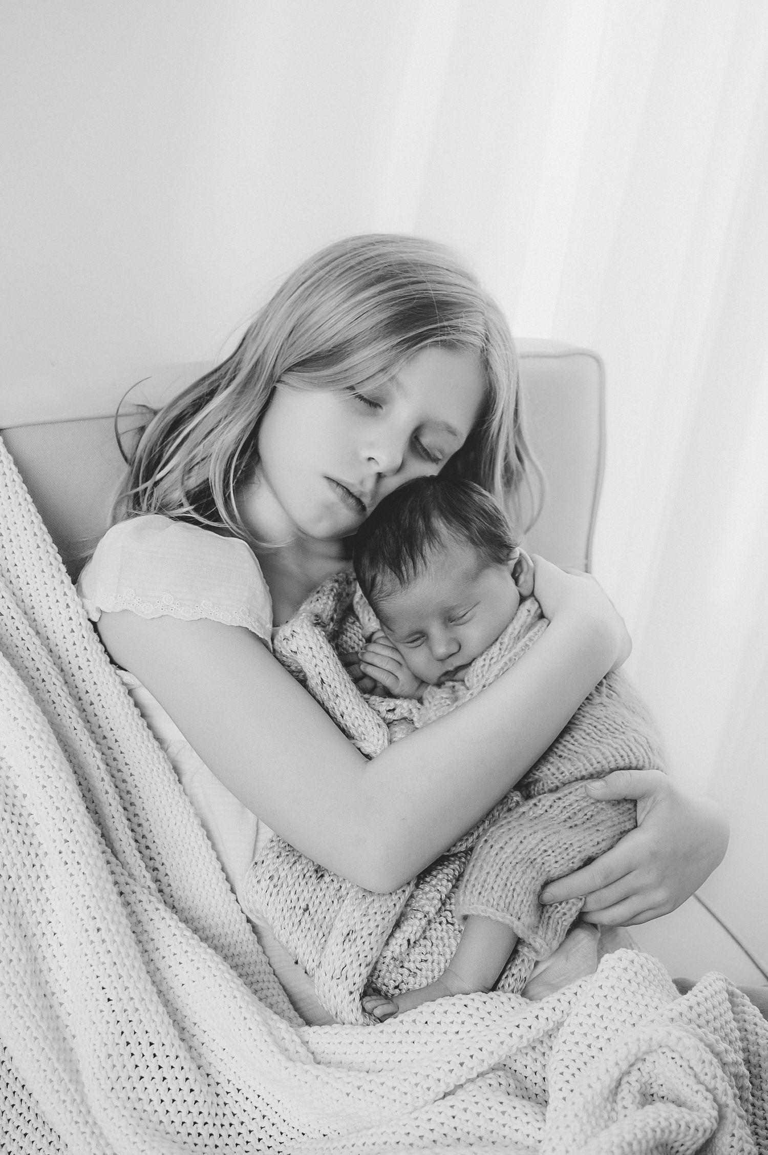 Black and white photo of a young girl tenderly holding a sleeping newborn baby wrapped in a knit blanket.