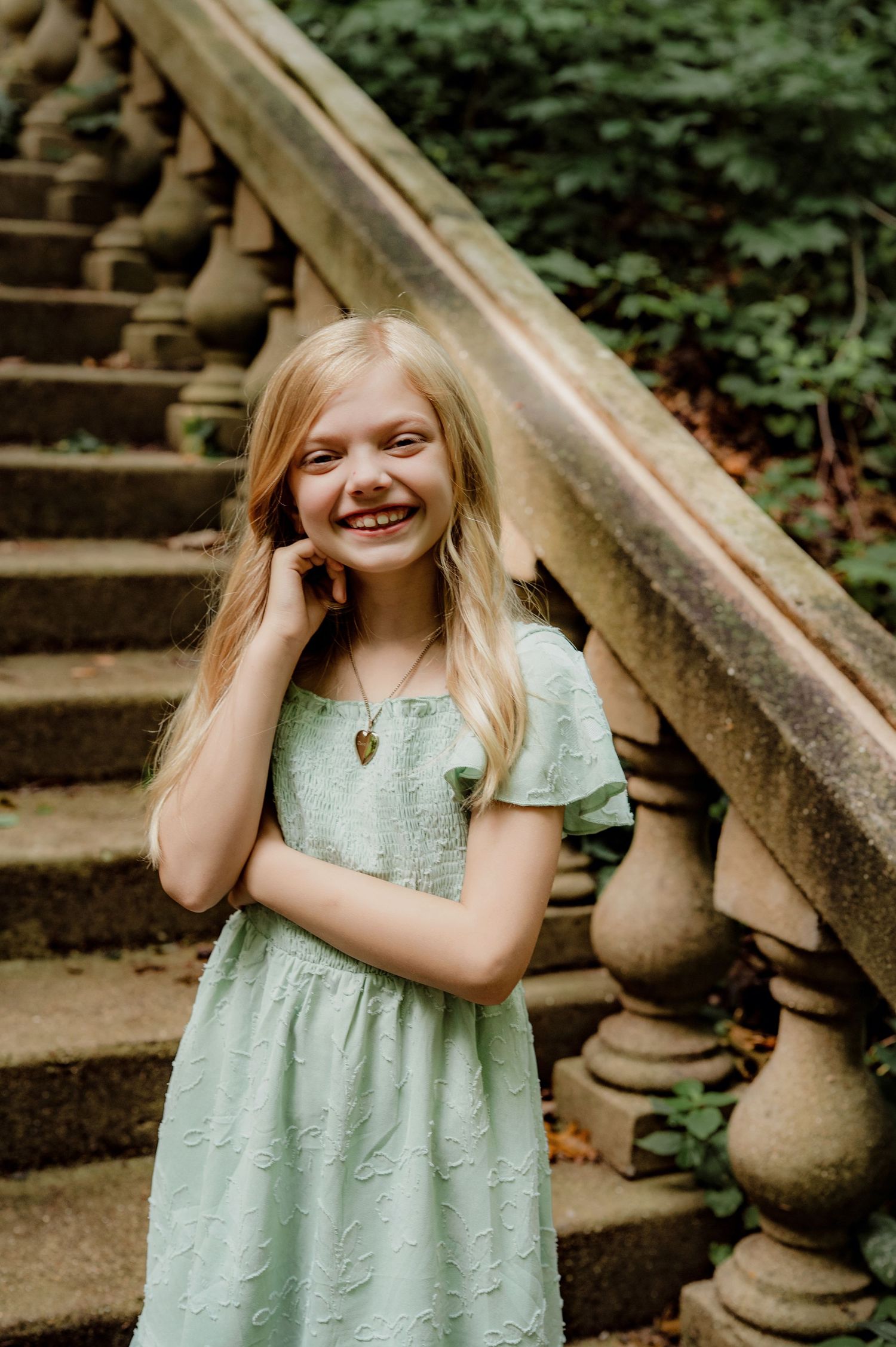 A smiling blonde girl in a mint green dress stands by stone stairs surrounded by lush greenery.